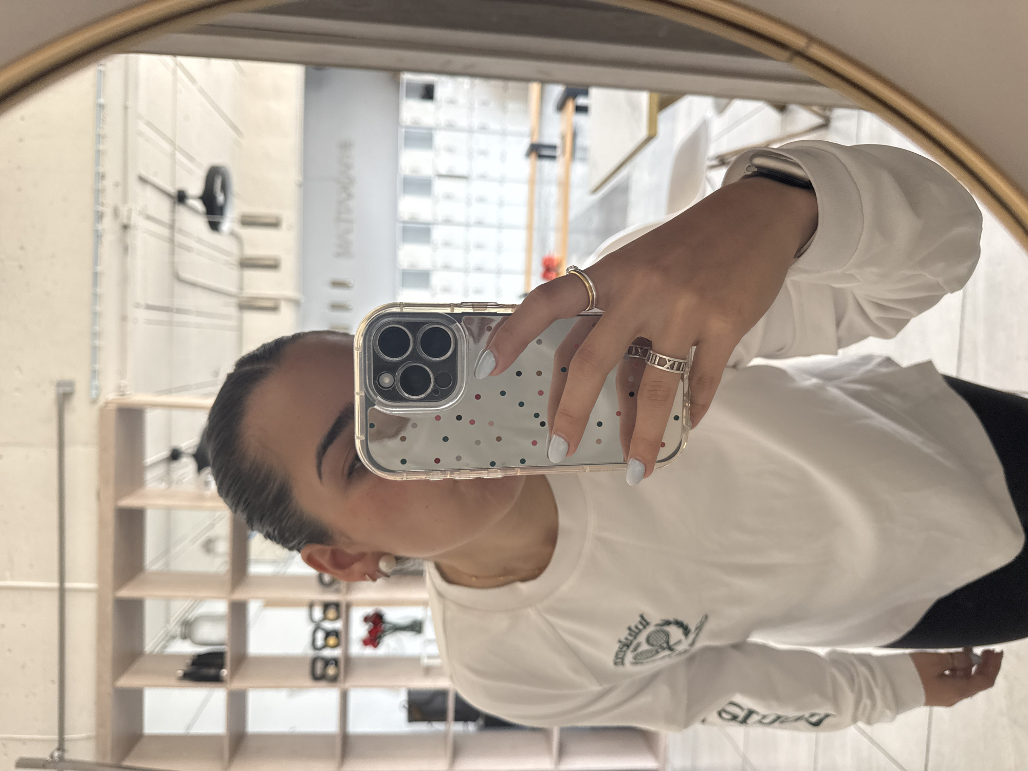 The image shows a person taking a selfie in a mirror. The person has light skin, dark hair pulled back, and is wearing a white long-sleeved shirt and black pants. They are holding a phone with a clear case decorated with small colorful dots, and wearing a gold ring and a silver ring with Roman numerals. The background is a room with white walls, tiled floors, and shelves displaying various items. A vertical sign with text is visible in the background. Based on available visual information and context, it is not possible to confirm the specific location in Puebla, Mexico.