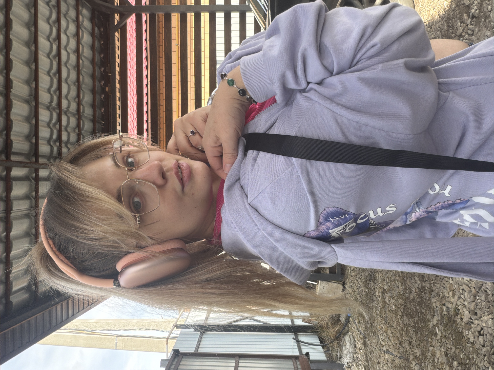 A young woman wearing eyeglasses and headphones is the focal point of the image. She is positioned outside near a structure with vertical slats of brown, pink, and metal, possibly a fence or an awning. Her attire consists of a lilac-colored sweatshirt. The surface beneath her appears to be gravel. The location is likely in or near the city of Большая Шильна, Russia.