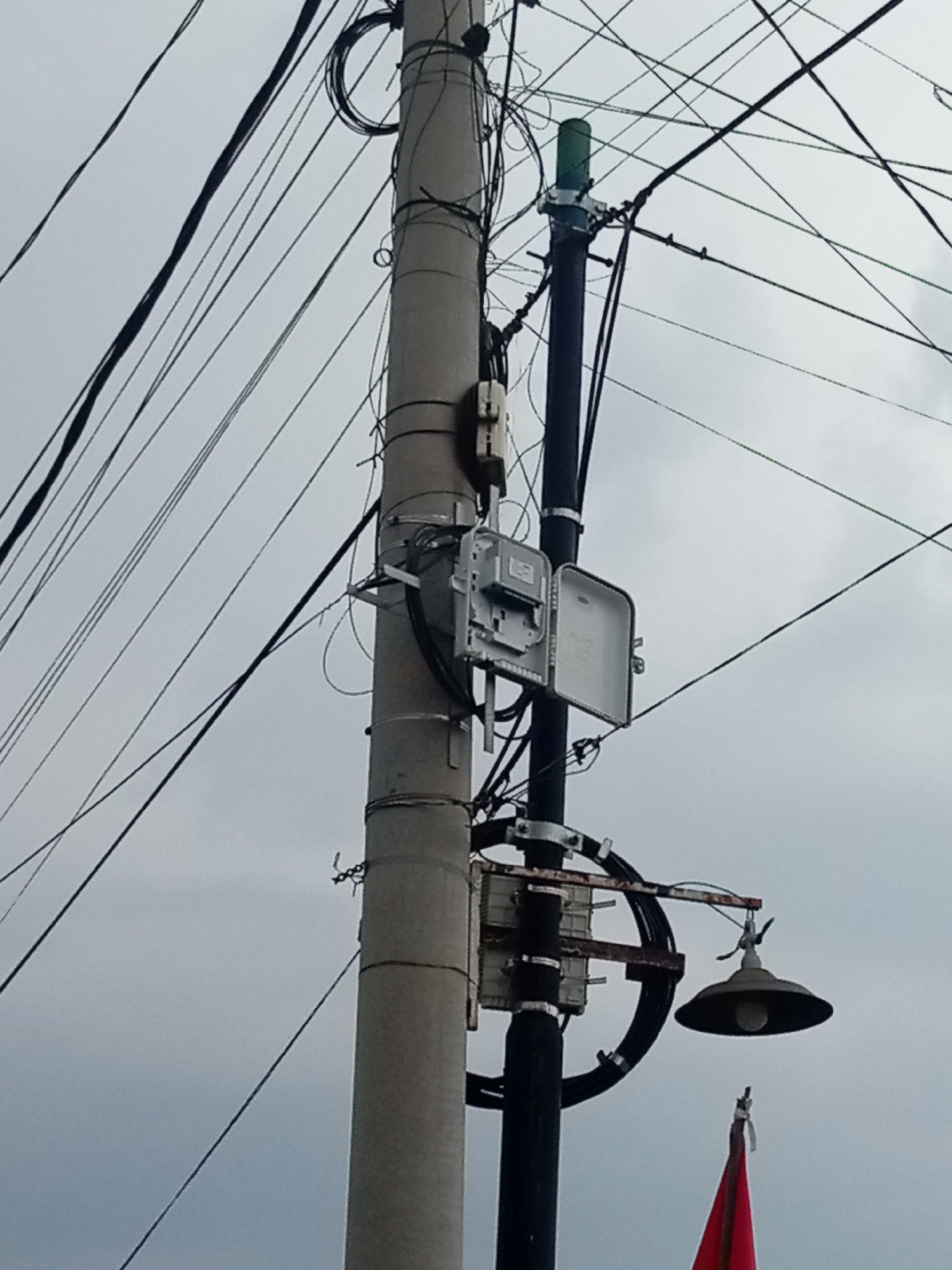 This is a low-angle, medium shot of a utility pole in Batam, Indonesia, against a cloudy sky. The pole is made of concrete and is covered in a tangle of black electrical wires. Attached to the pole are several pieces of equipment, including an open gray electrical box with visible internal components, a green cylindrical object at the top, and a street lamp with a conical shade. A small red flag is visible at the bottom of the frame. The scene appears to be outdoors, likely on a street, given the presence of the utility pole and street lamp. The overall impression is one of urban infrastructure and electrical distribution.