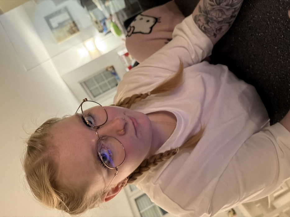 A woman with blonde hair in pigtails peers upwards, wearing round glasses and a white long-sleeved shirt. Her nose is pierced, and a tattoo is visible on her arm. She is partially sitting on a dark colored sofa; a pink pillow with a Hello Kitty print is also visible. The background shows a glimpse of a room with neutral walls, a window, and shelves. The perspective is from a lower angle looking up at the woman.
