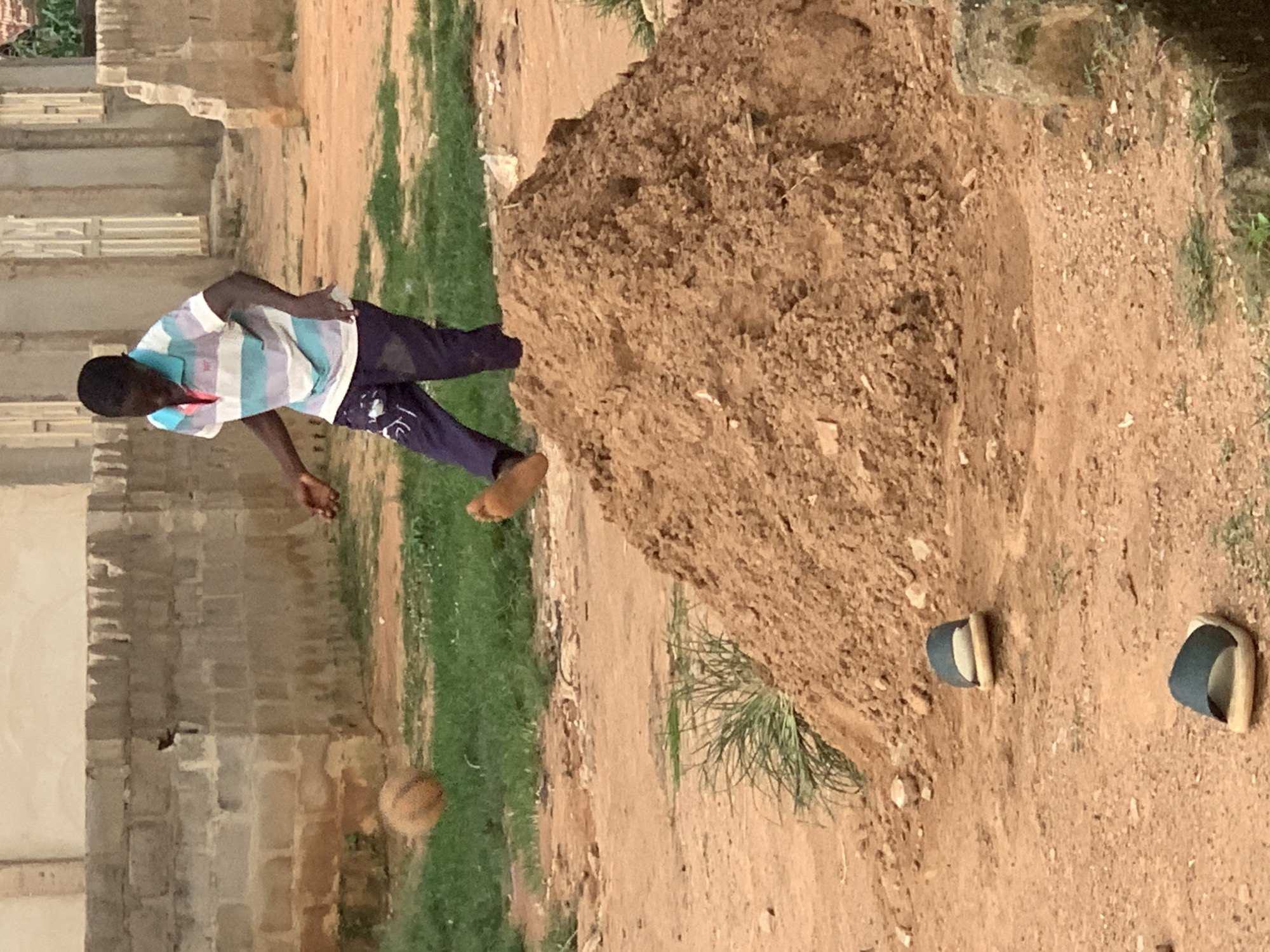 In Sabon Tasha, Nigeria, a young boy is seen playing with a ball on a dirt field, with a pile of dirt in the foreground and buildings in the background. The boy, wearing a striped t-shirt and dark pants, is in mid-action, with his right leg extended as if kicking the ball, which is airborne to his left. Two pairs of open-toed sandals are visible on the ground in the foreground. The environment appears to be an unpaved area with scattered grass and construction elements, suggesting a developing urban or semi-urban setting.