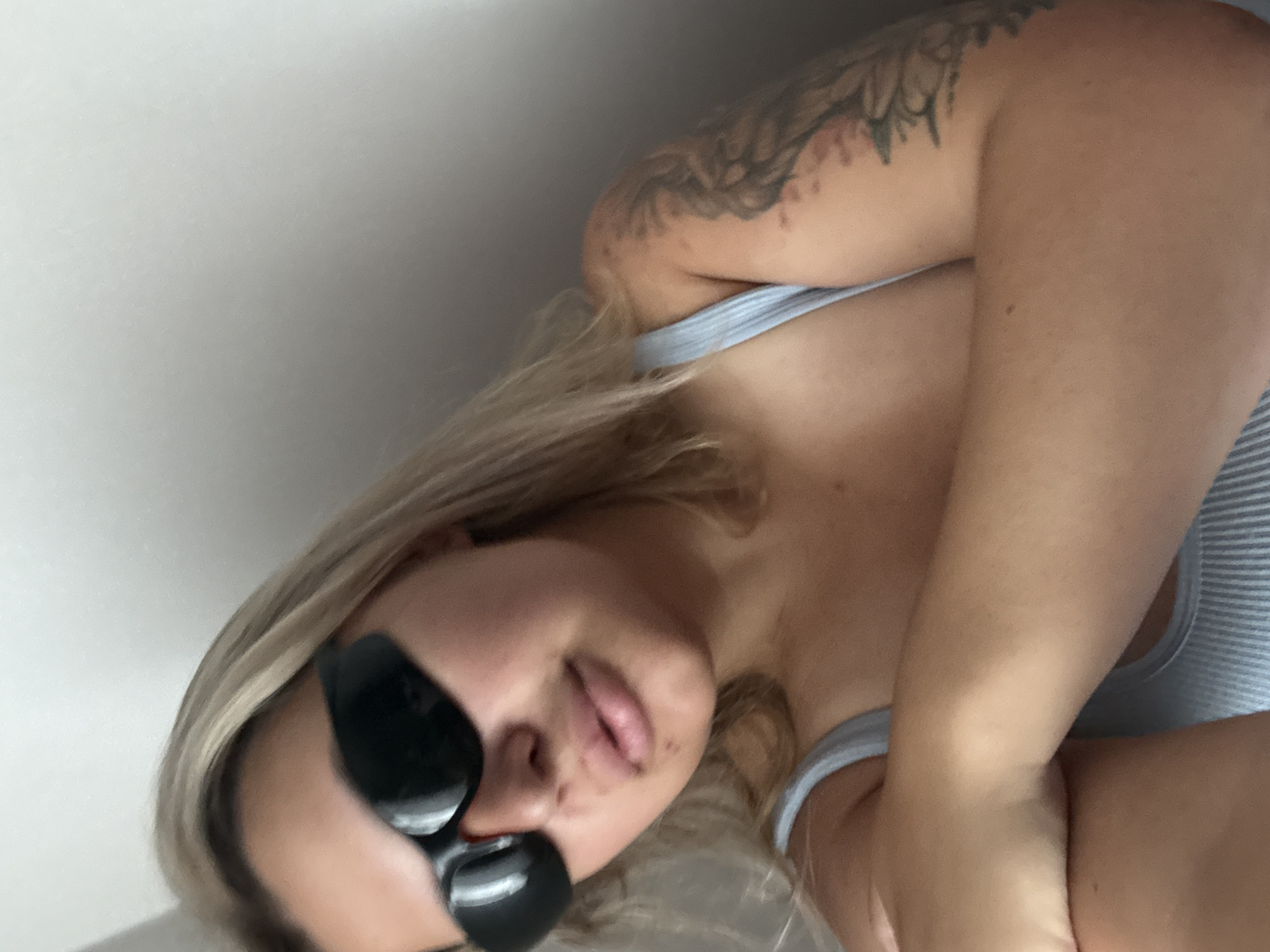 A young woman with blonde hair and sunglasses is seen from the chest up, looking towards the camera. She is wearing a light blue tank top. Her left arm is crossed over her chest, obscuring part of her torso. A tattoo is visible on her right upper arm. The background is a plain, light-colored wall. The image appears to be a selfie taken indoors. There is no indication of the location being Khabarovsk, Russia, or any specific city.