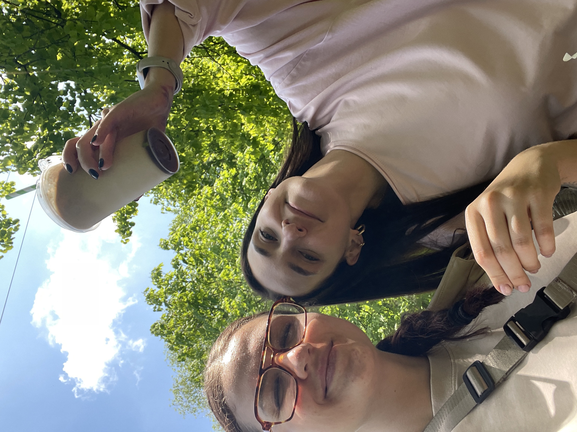Two people are in the foreground, taking a selfie outdoors. The bottom person is wearing glasses and has their hair in a ponytail. The top person is holding a drink in their left hand, with trees and blue sky in the background. The location cannot be determined.