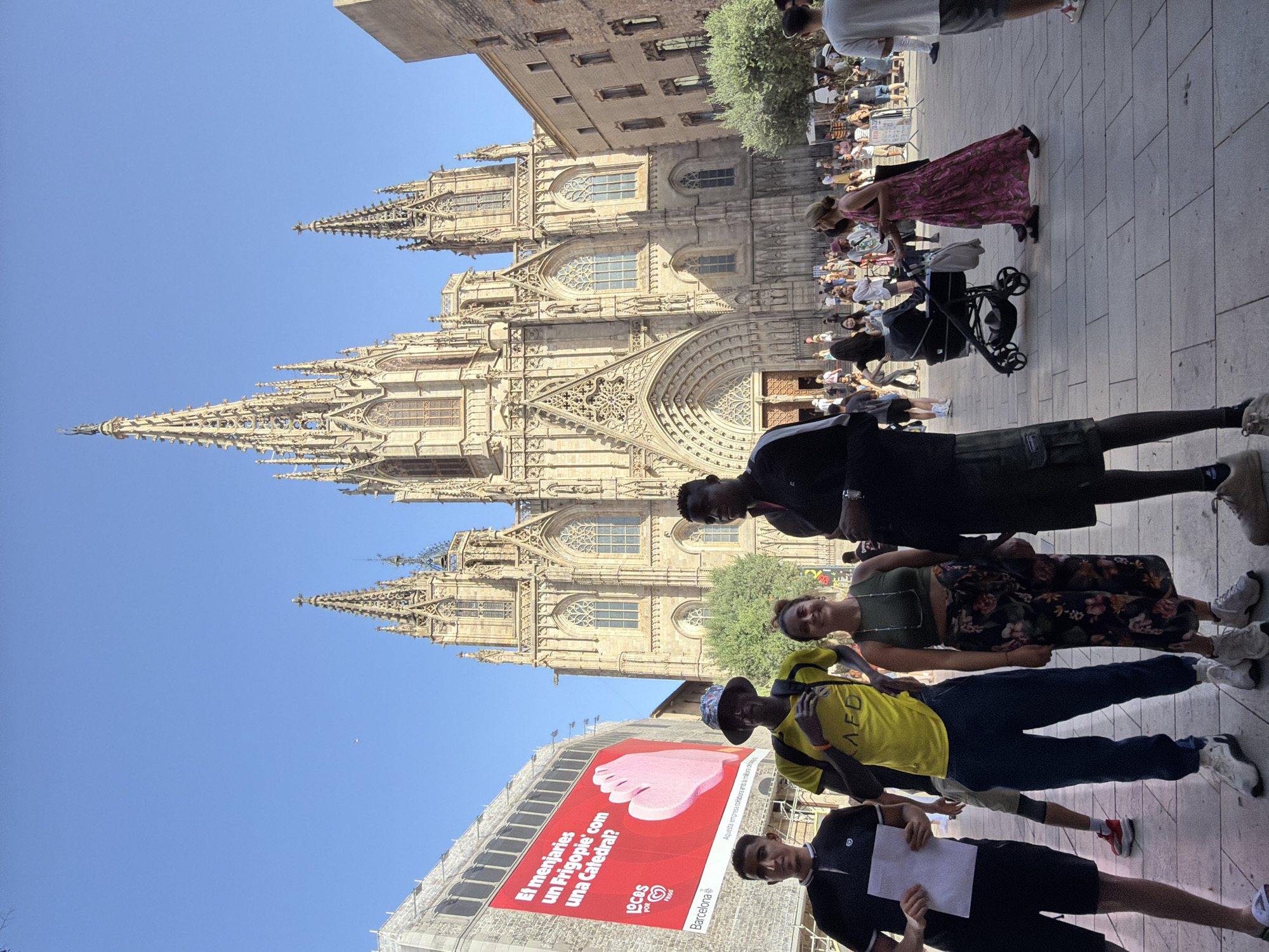 The image captures a sunny day in Barcelona, Spain, with the Barcelona Cathedral prominently featured in the background. The Gothic architecture of the cathedral dominates the scene, with its intricate spires and detailed facade bathed in sunlight. In the foreground, a group of four individuals stands on a paved plaza. Two men are on the left, one wearing a black polo shirt and holding a white paper, the other in a bright yellow t-shirt with "AED" written on it and dark pants. A woman in a green top and a floral-patterned skirt stands between the two men. A fourth individual, a man in a black jacket, stands to the right of the woman.
Beyond the central group, the plaza is populated with numerous people, suggesting a tourist area or public gathering. A woman pushing a stroller walks across the plaza, and others are seen milling about, some possibly tourists or locals enjoying the day. To the left of the cathedral, a large red billboard advertising "LOCAS por Frigo" is visible, with a pink depiction of what appears to be an ice cream cone. Trees provide greenery within the urban landscape. The sky is clear and blue, indicative of good weather.