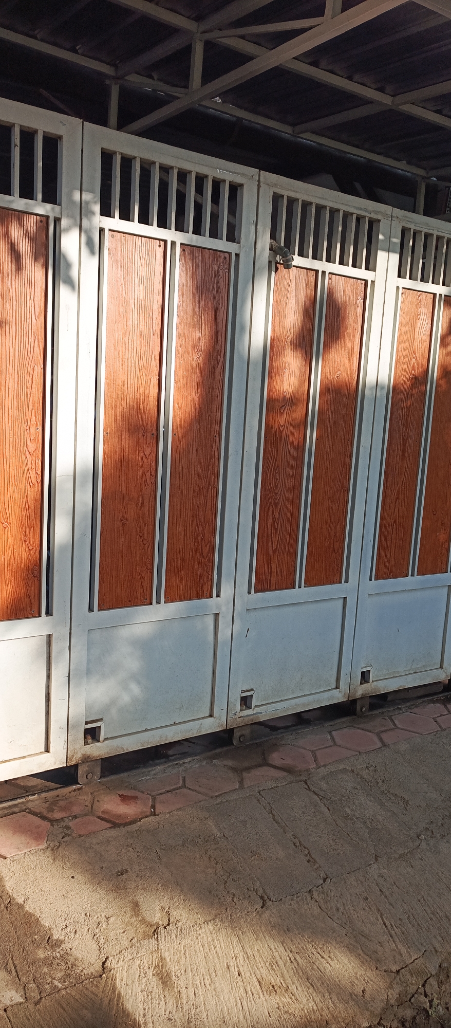 The image shows a closed gate composed of several vertical panels. The upper portion of the gate has a barred design, while the lower section features vertical wooden-patterned planks within a white frame. The gate appears to be made of metal. The ground in front of the gate is paved with hexagonal bricks and concrete. Sunlight casts shadows on the gate and the ground. A metal structure with a dark roof is visible above the gate. The scene is located on a paved surface that appears to be part of a residential or commercial property in Jatisari, Indonesia.