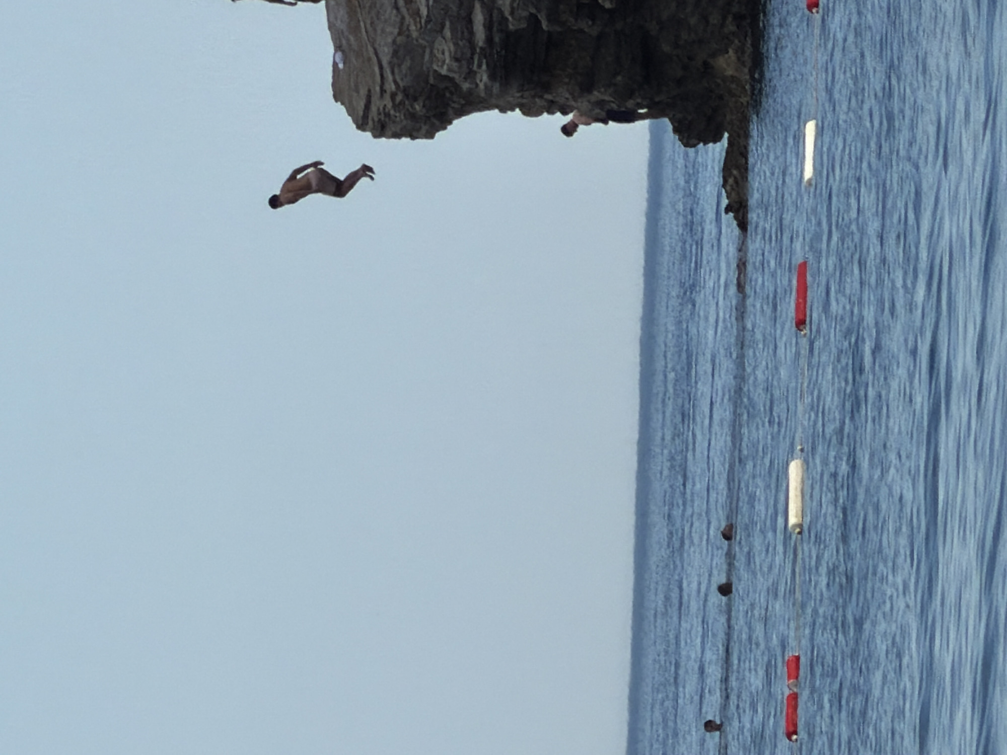 A man is captured mid-air, leaping from a rocky cliff into the blue sea, with another man standing on the cliff edge above him. In the water below, several people are swimming, and buoys are visible, with two red buoys and one white buoy in the foreground. The scene appears to be a popular spot for cliff diving, likely in Budva, Montenegro, known for its rocky coastlines and clear waters. The sky is a pale blue with no clouds.