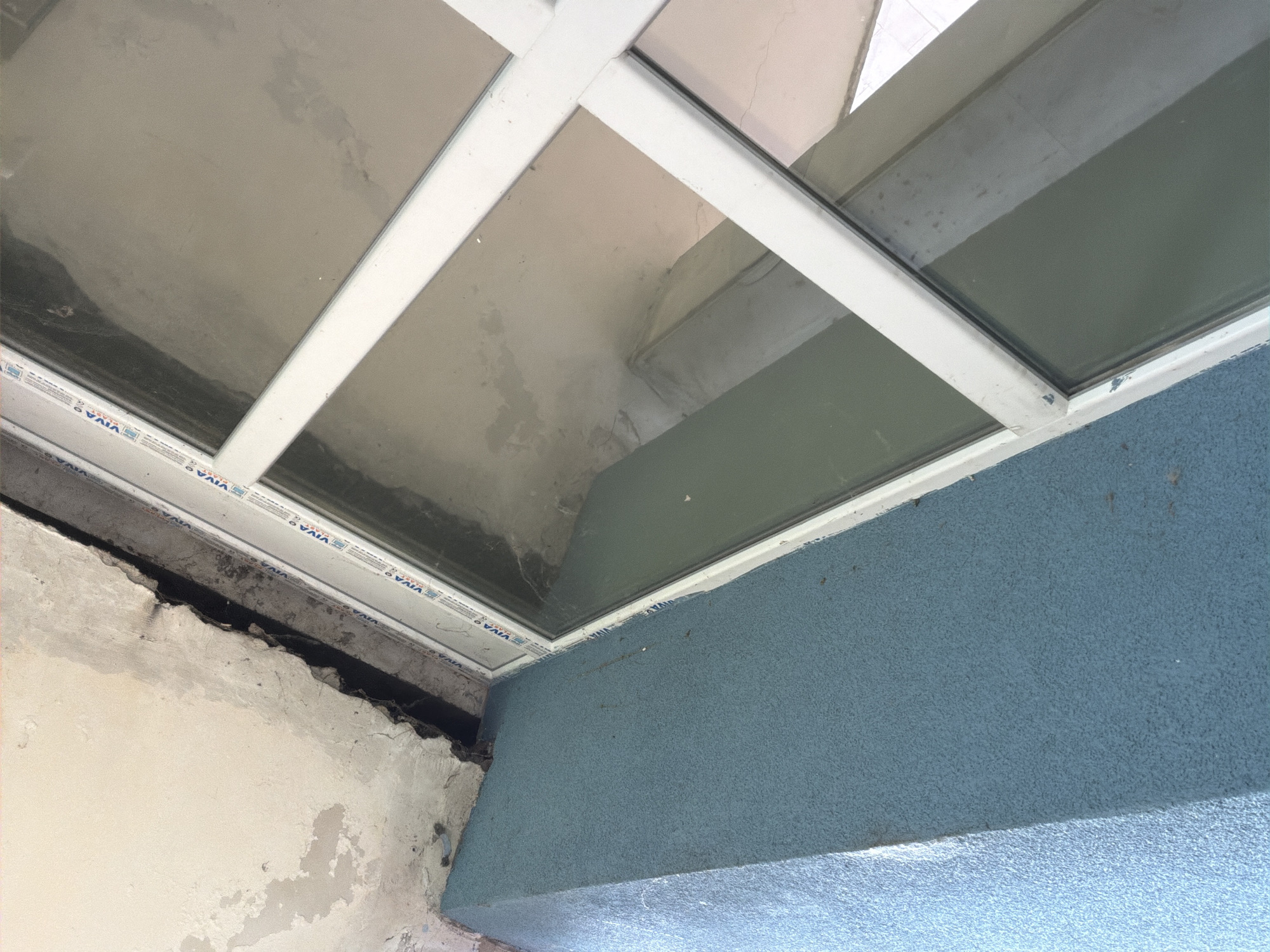 An interior view showing a white-framed skylight above a wall, possibly in Dolna Mitropolia, Bulgaria. The wall is textured blue, while the adjacent wall is peeling and grey. Visible above is the window and parts of the ceiling in varying shades of gray.