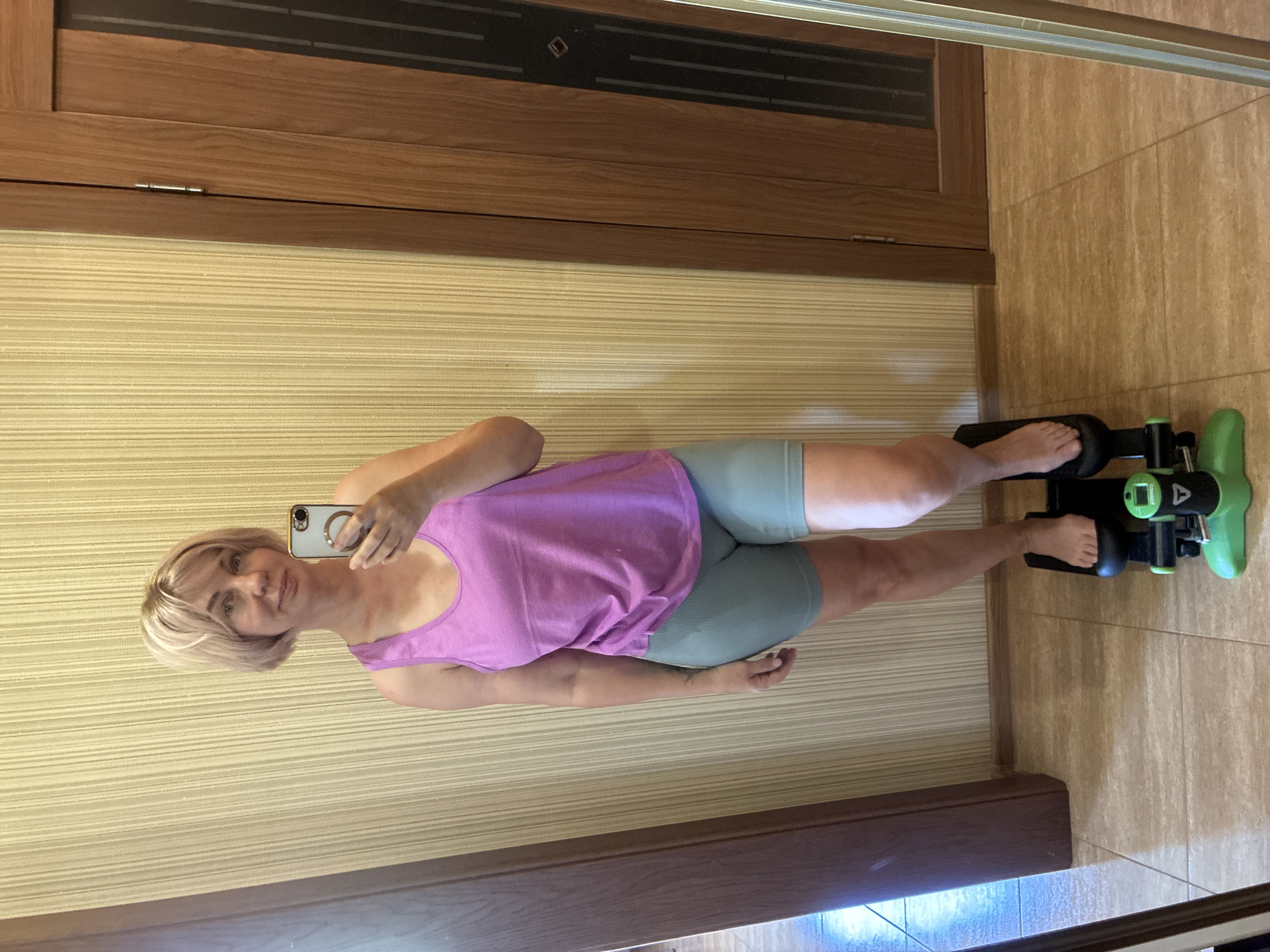 An individual is depicted taking a mirror selfie indoors. The subject is a woman with fair skin and short, light-colored hair, appearing to be in her 30s or 40s. She is wearing a purple sleeveless tank top and light green, ribbed biker shorts. Her bare feet are positioned on a green and black compact stepper fitness machine, branded "ZOOMGEO," which rests on a light brown tiled floor. She holds a silver smartphone with a gold ring stand in her right hand, directed towards the mirror. The background features a light beige wall with a vertical textured pattern. To the right, a wooden door with a dark vertical panel and a visible hinge is present. A partial reflection of a dark wooden structure is visible on the left side of the mirror's frame.