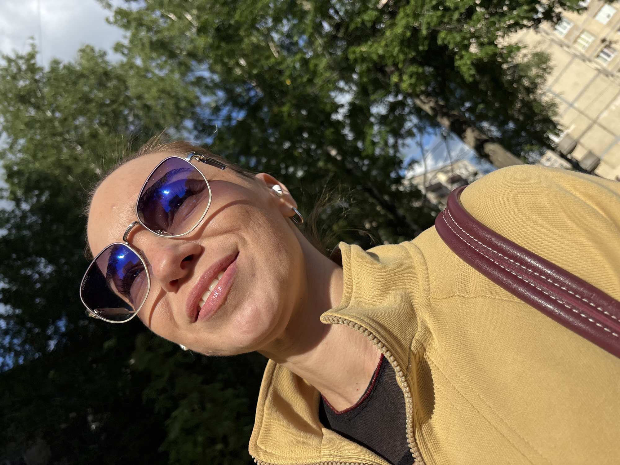 A close-up, low-angle shot captures a woman looking up towards the camera, her face partially obscured by sunglasses. She is wearing a yellow zip-up jacket over a black top. Her lips are slightly parted in a smile, revealing her teeth. The sunglasses have reflective lenses, showing a blue sky and some greenery. She is also wearing discreet white earbuds and a dark red strap from a bag crosses her shoulder.
The background consists of out-of-focus green trees against a bright blue sky, suggesting an outdoor setting during the day, likely with good weather. Hints of buildings are visible in the upper right portion of the image. There is some visible text on the side of her sunglasses, but it is not legible. The overall impression is of a casual, sunny moment.