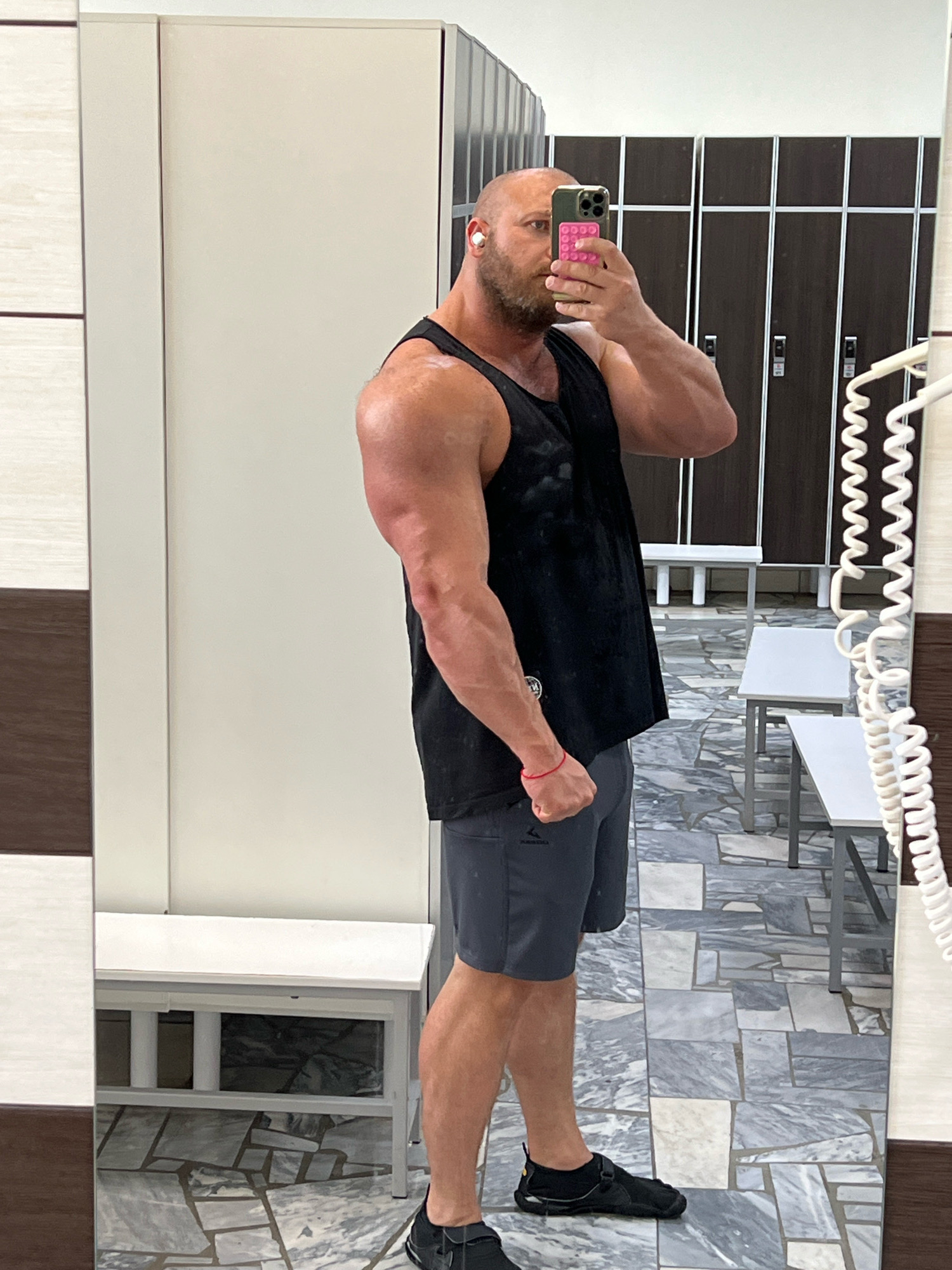 A bald man with a beard and earbuds is taking a mirror selfie in a gym locker room. He is wearing a black tank top and grey shorts, and has a red bracelet on his left wrist. The locker room features rows of dark brown lockers, white benches, and a tiled floor. The man's phone case has a pink pop socket. This scene is located in Chaikovsky, Russia.