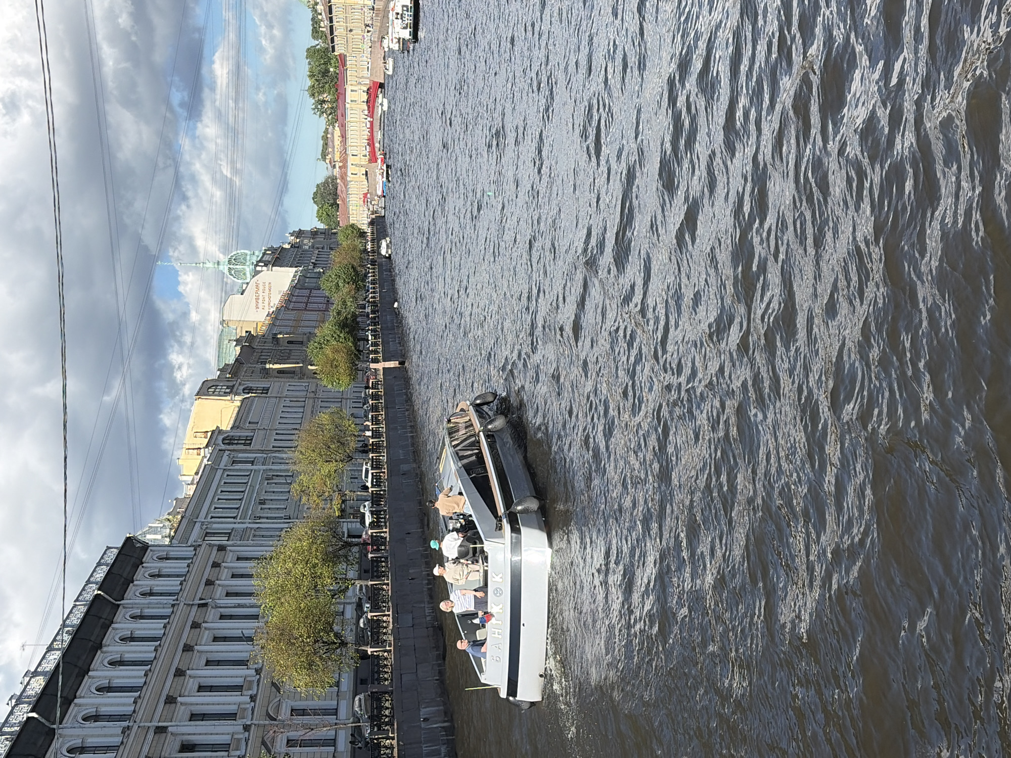 This media file depicts a boat tour on a river in Saint Petersburg, Russia. The scene is a picturesque urban landscape with historic buildings lining the riverbank. The boat, a sleek, modern design with a cabin and open seating area, carries several passengers. The people on the boat appear relaxed and engaged in the tour, suggesting a leisurely daytime activity.
The time of day appears to be daytime, with the sun casting shadows and illuminating the water. The weather seems pleasant, with a mix of clouds and sunshine in the sky. The buildings in the background showcase classic European architecture, typical of Saint Petersburg, with ornate facades and tall windows. The river itself is calm, with gentle ripples reflecting the sky and surrounding structures. Trees line the riverbank, adding a touch of greenery to the urban environment.
Notable details include the text visible on the side of the boat, which reads "БАЙКОК" (Baikok). The overall mood is serene and tourist-oriented, characteristic of a city that attracts visitors with its historical significance and scenic waterways. The presence of overhead wires suggests the proximity of infrastructure, possibly a bridge or streetcar lines, further grounding the scene in an urban setting. The composition offers a view from a slightly elevated perspective, looking down onto the river and the boat.