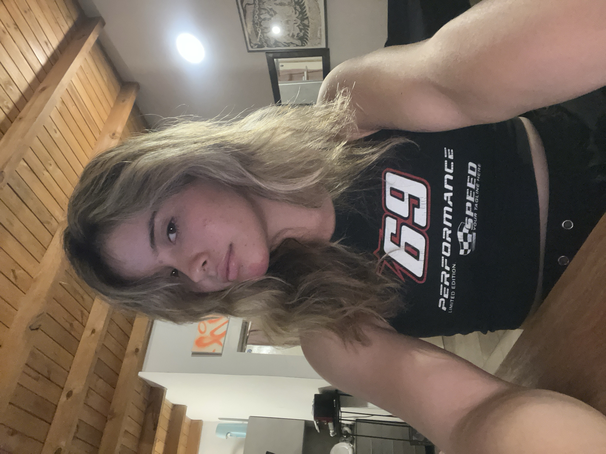 A woman is indoors, taking a selfie. She is wearing a black tank top with "69" and "Performance Speed" graphics. The location appears to be a room with wood paneling on the ceiling. There are framed pictures on the wall. A kitchen area with a sink and other items is visible in the background. The location is Sumalao, Argentina.