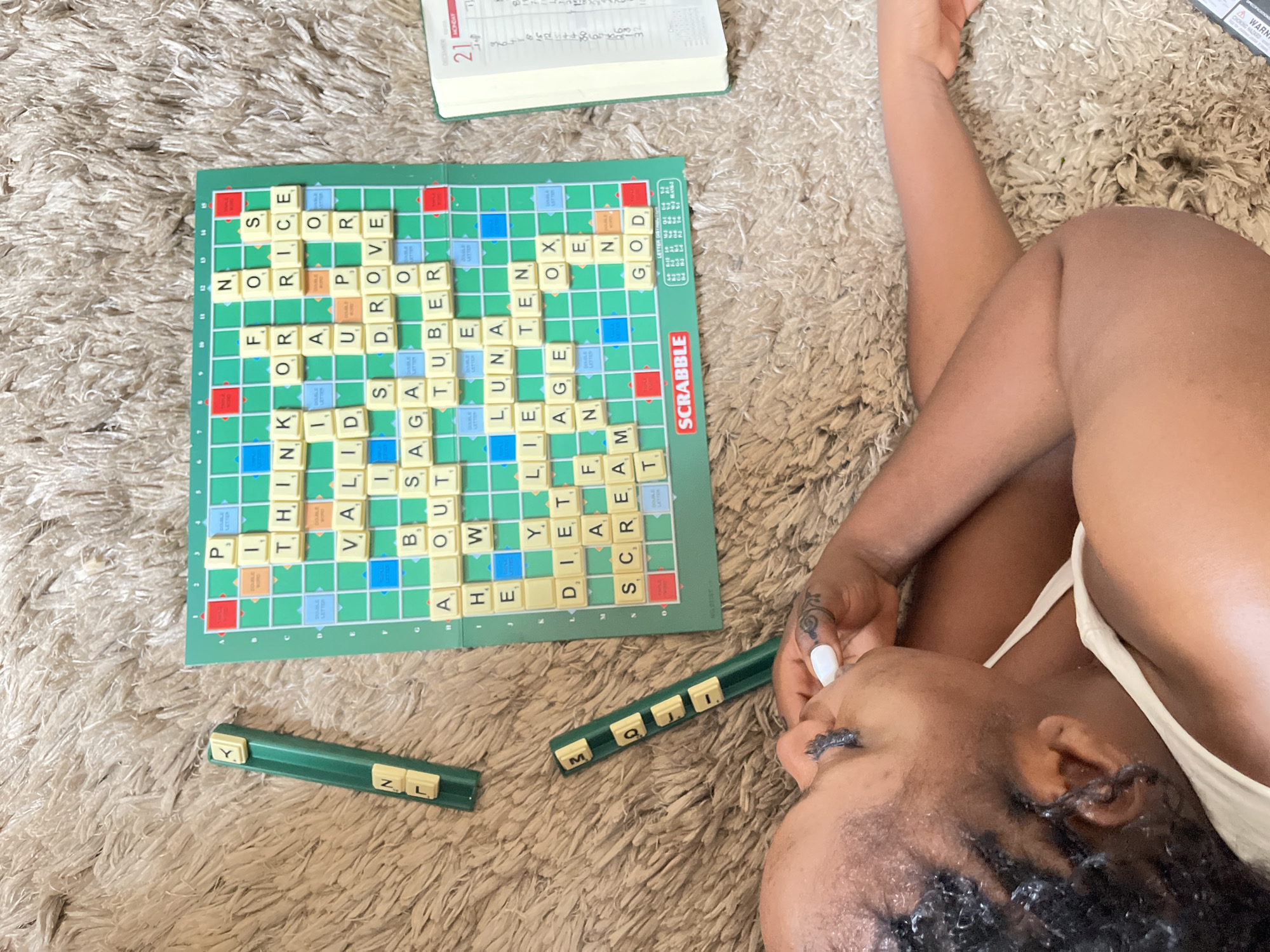 An overhead view captures a person relaxing on a fluffy, light-colored rug, engaged in a game of Scrabble in an indoor setting in Karmajiji, Nigeria. The individual, with dark skin and braided hair, is seen from the upper body, lying on their side with one arm bent near their head. They are wearing a light-colored top, and their eyes appear closed or looking downwards, suggesting a moment of relaxation or contemplation.
The main subject is a Scrabble board, densely filled with played words. Visible words on the board include "THINK," "VALID," "SAGA," "OUT," "HE," "LUNA," "LIE," "DIET," "SCREAM," "FOR," "RICE," "UP," "DROVE," "TUBER," "AGE," "TEN," "OX," and "GOD," among others. The board also displays the game's title "SCRABBLE" and common bonus square labels such as "DOUBLE LETTER," "TRIPLE LETTER," "DOUBLE WORD," and "TRIPLE WORD."
Two green Scrabble tile racks are positioned on the rug. One rack holds the letters 'Y', 'N', and 'L', while the other contains 'M', 'Q', 'J', and 'I'.
In the upper part of the image, an open planner or diary is visible, indicating "DECEMBER," "MONDAY," and the date "21," with some handwritten notes. A small part of a warning label, featuring "CHOKING HAZARD" and "WARNING," is also discernible. The person's hand shows white nail polish and a dark ring, with a faint tattoo-like design on their skin. The scene conveys a peaceful and leisurely activity.