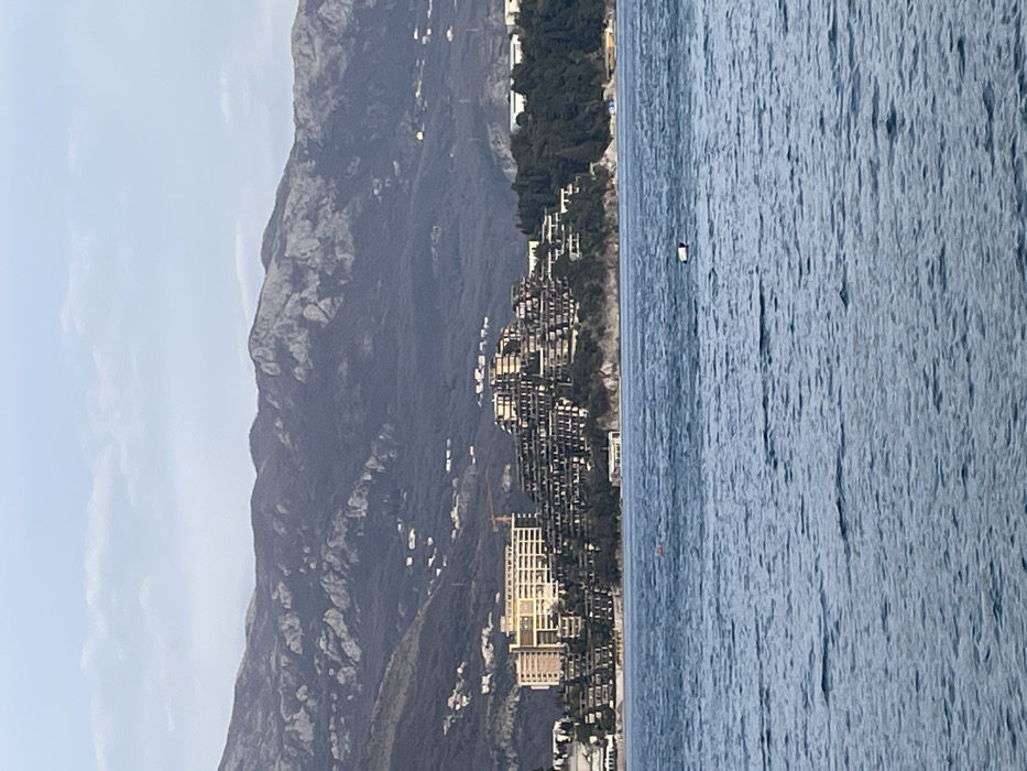 The image displays a panoramic view of the coastline near Budva, Montenegro, observed from the sea. The foreground consists of rippled, blue-grey water, containing a white and black rectangular object, possibly a buoy, floating slightly right of center, and a smaller orange cylindrical object to its left.
The midground features a prominent landmass, identified as the Zavala Peninsula and parts of the Bečići coastline. On the left section of this landmass, a large, multi-story, light-colored hotel complex with numerous balconies is visible, situated near the shore. A yellow construction crane is present behind this building. To its right, covering the elevated slope of the peninsula, are multiple dark-colored, terraced or stepped building structures with visible balconies, consistent with the architecture of the Dukley Gardens resort. Patches of green vegetation and trees are interspersed among these buildings and along the shoreline.
The background is dominated by a range of rugged, dark mountains exhibiting rocky outcrops and sparse vegetation. Scattered small white buildings are visible on the lower slopes of these mountains. The sky above is pale blue with minimal, faint white cloud formations, indicating daytime conditions. No individuals are depicted in the scene.