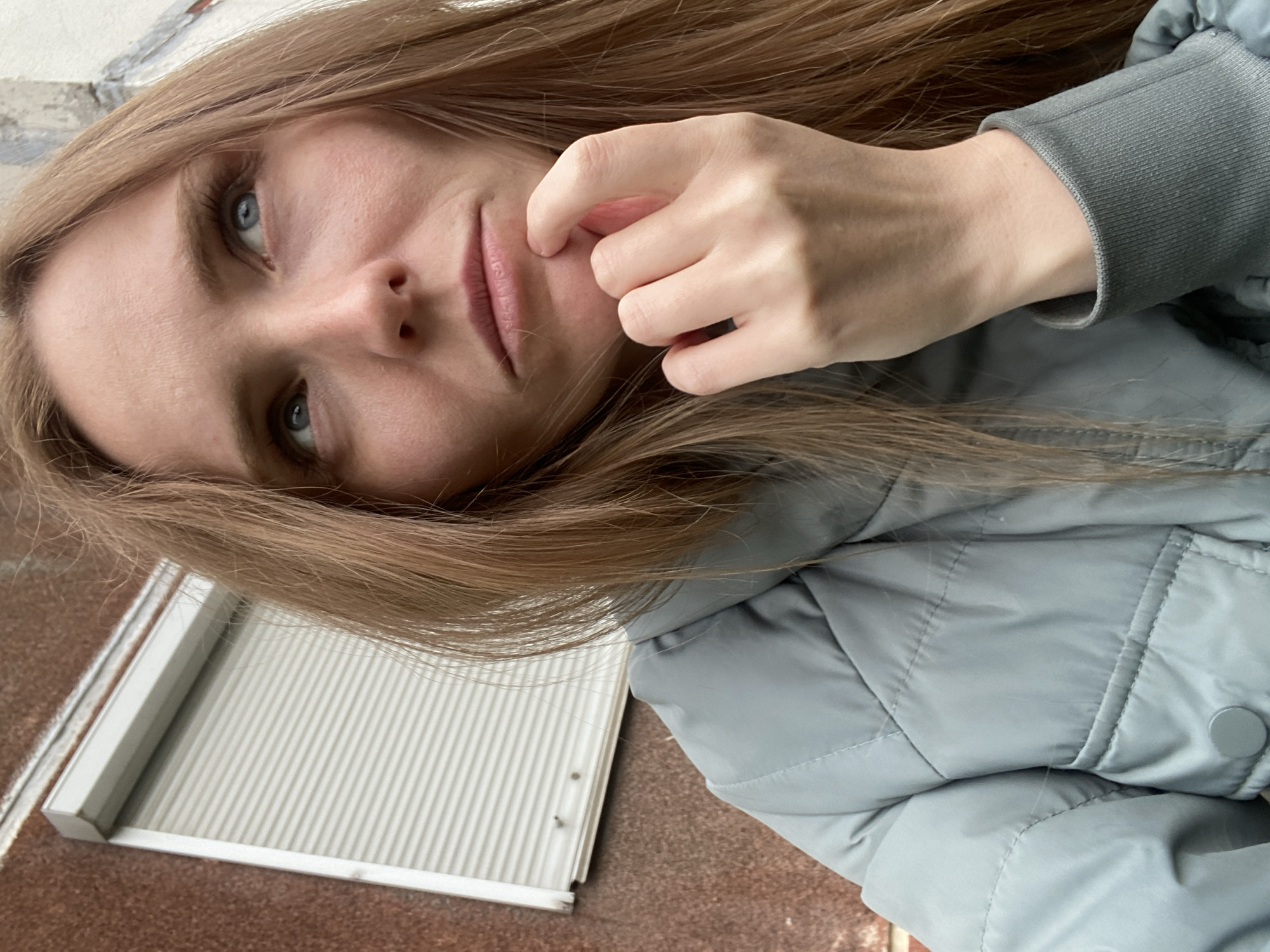 A young Caucasian woman with long, straight, light brown hair is captured in a close-up, waist-up shot, looking upwards and to the right. She is wearing a light blue puffer jacket over a gray sweatshirt. Her right hand is resting on her chin, with her index finger touching her lower lip, suggesting a pensive or contemplative mood. The background is out of focus, showing a textured, reddish-brown wall on the left and a portion of a building with a light-colored, ribbed shutter on the right. The overall lighting appears to be overcast. The image was taken in the city of Yuzhno-Sakhalinsk, Russia.