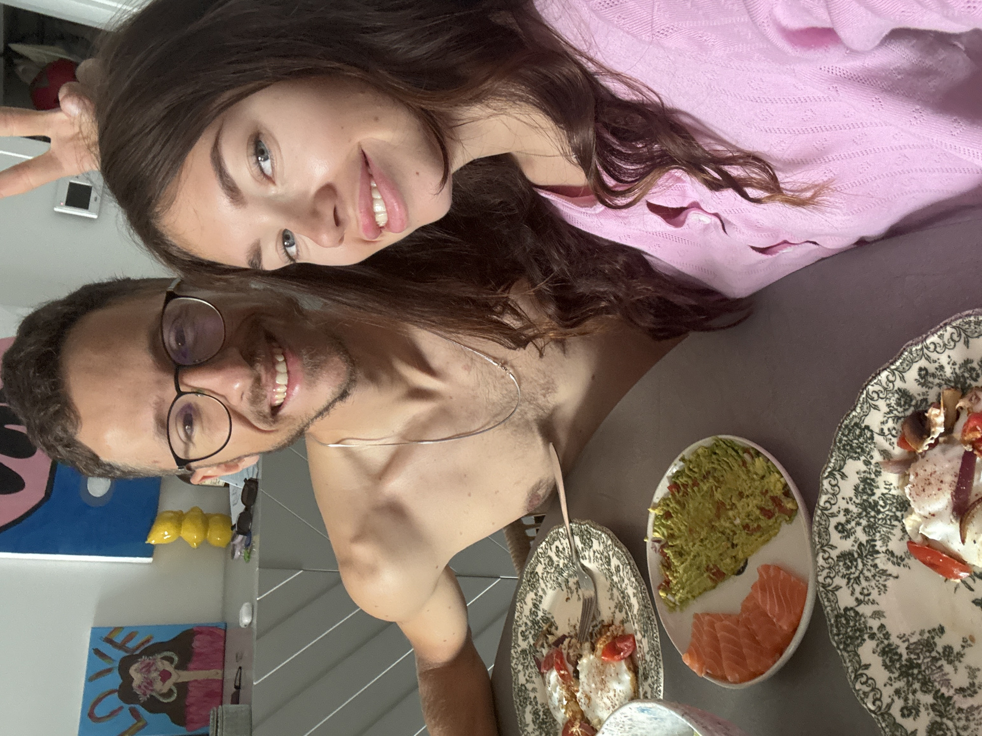 A selfie depicts a man and a woman indoors in Tel-Aviv, Israel, posing at a table with plates of food. The woman, positioned above the man, wears a pink top and has brown hair. The man, shirtless and wearing glasses, is next to her. The table holds three plates with varying foods including scrambled eggs, avocado, and sliced salmon. The setting appears to be a home dining area. A painting and yellow decorative items are visible in the background.