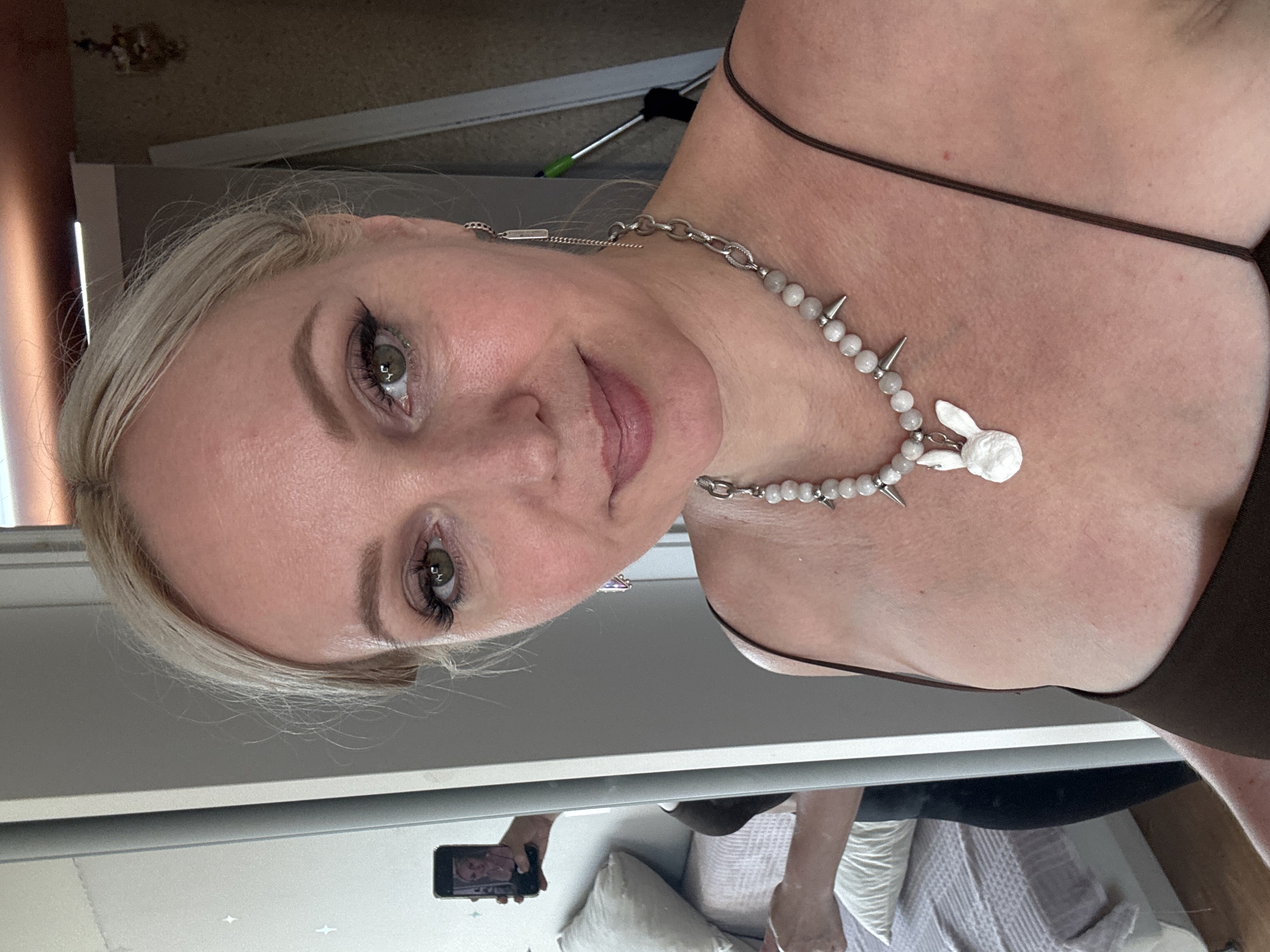 A woman with blonde hair and green eyes is taking a mirror selfie. She is wearing a brown camisole and a necklace with a bunny pendant. The background shows a bedroom with a bed and a closet. The image does not contain any elements that would allow for the identification of its location in Komsomolsk-on-Amur, Russia.
