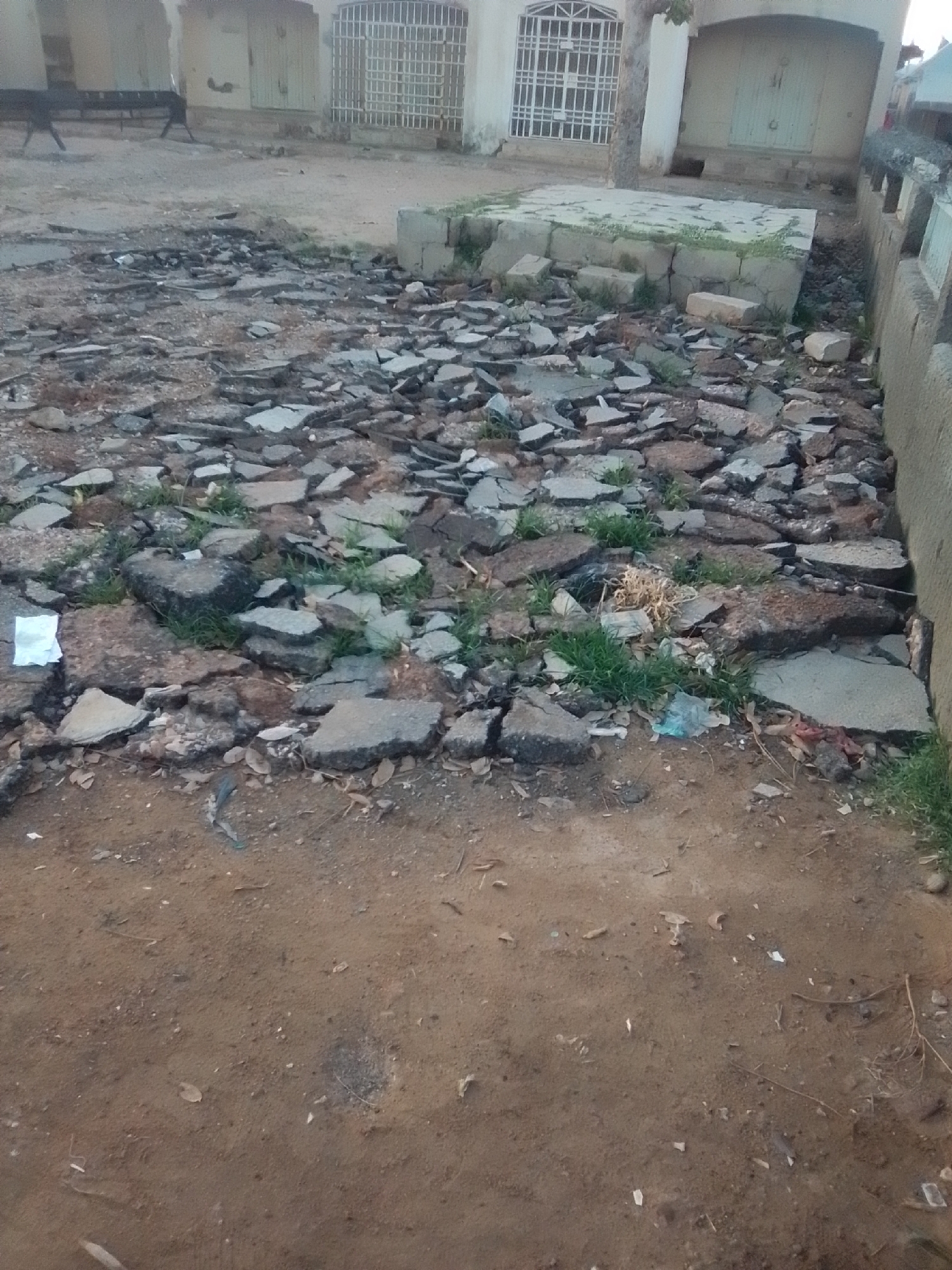 The image shows a street in Gombe, Nigeria. The street is made of asphalt and is broken into pieces. There is also some debris on the street, including rocks, grass, and trash. The buildings on either side of the street are made of concrete and are painted white. There is a fence in front of one of the buildings. The sky is overcast.