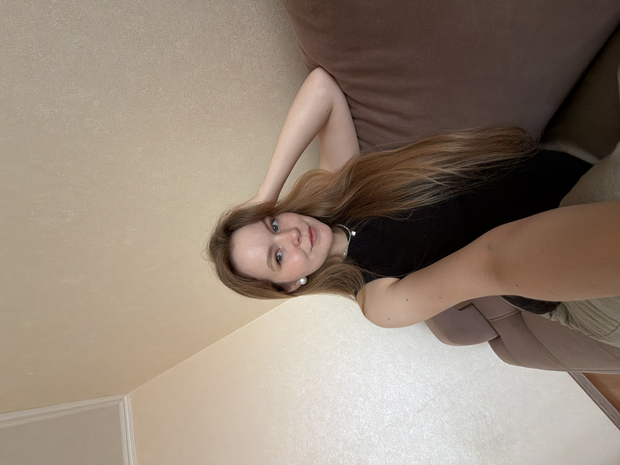 A young woman with long, light brown hair is sitting on a brown couch in what appears to be a residential setting. She is wearing a black sleeveless top and a necklace with a heart-shaped pendant. She also has large pearl stud earrings. Her right arm is resting on the back of the couch, and her left hand is supporting her head. The background consists of a beige-colored wall. The lighting suggests an indoor environment, likely during the daytime. There is no discernible location in the city of Kazan, Russia, or any specific context provided by the image.