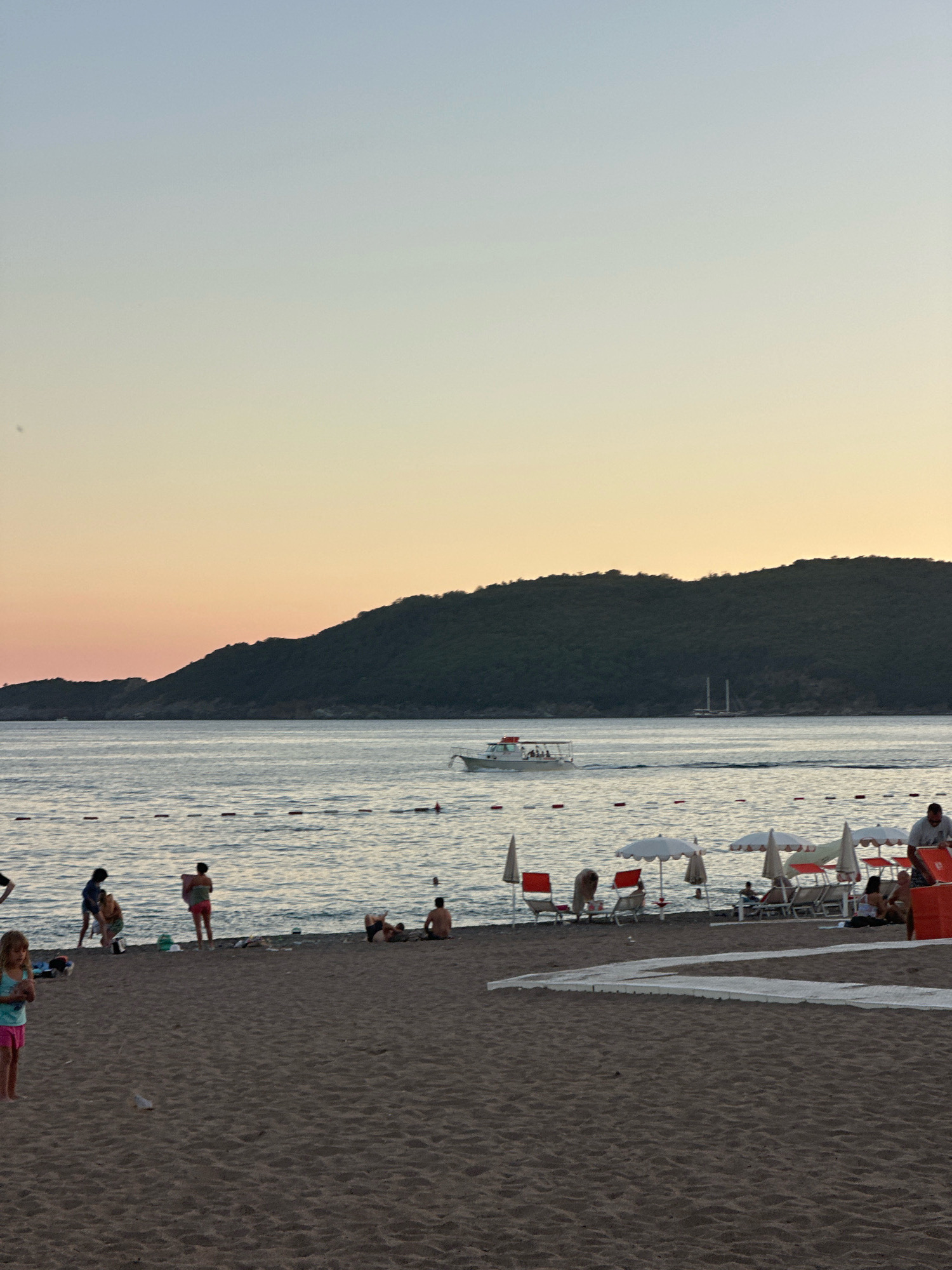 In Boreti, Montenegro, at dusk, a beach scene unfolds along the Adriatic Sea. The sandy shore is populated with scattered individuals, some engaged in conversation, others relaxing on loungers beneath umbrellas. A few children play near the water's edge. In the middle distance, a small boat navigates the calm sea, leaving a faint wake. Further out, a large sailboat is visible on the horizon, silhouetted against the fading light. The background is dominated by a dark, tree-covered mountain that slopes down to the water. The sky transitions from a pale blue at the top to a soft orange and pink hue near the horizon, indicating the close of day. A white pathway or mat is laid out on the sand in the foreground, leading towards the water.