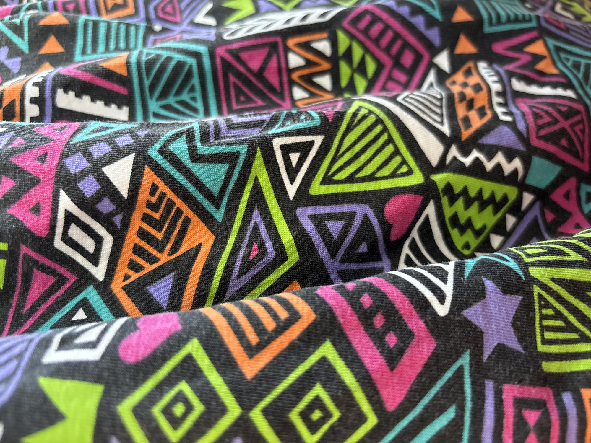 The image is a close-up, high-angle shot of a black fabric with a vibrant, geometric pattern. The pattern consists of various shapes, including triangles, squares, diamonds, and zigzags, rendered in bright neon colors such as pink, lime green, orange, purple, and turquoise. The fabric appears to be folded or draped, creating shadows and highlights that accentuate its texture and the folds of the material. There are no people or discernible environmental elements present, suggesting the image focuses solely on the textile design. The image does not provide any information about its location in Blagoveshchensk, Russia.