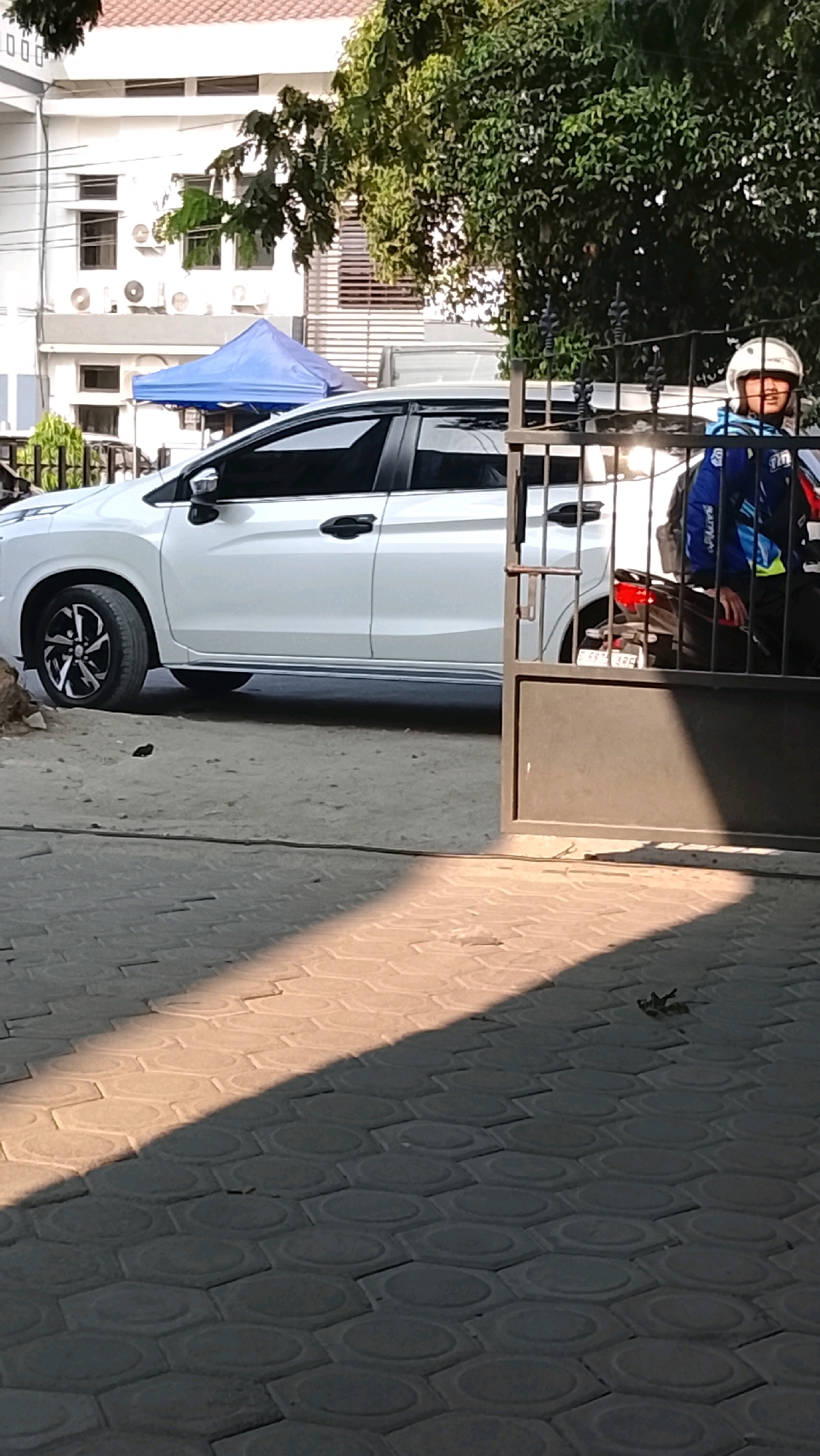 In Slawi, Indonesia, a white MPV is parked on a sandy lot in front of a building. The vehicle, with tinted windows and black and silver rims, is positioned to the left of a black metal gate. Behind the gate, a person wearing a white helmet and a blue and yellow jacket sits on a motorcycle, facing the MPV. The building behind the MPV has multiple windows and air conditioning units. A blue tent is set up in front of the building, to the left of the MPV. The foreground of the image is a paved area with a hexagonal pattern, illuminated by dappled sunlight creating strong shadows.