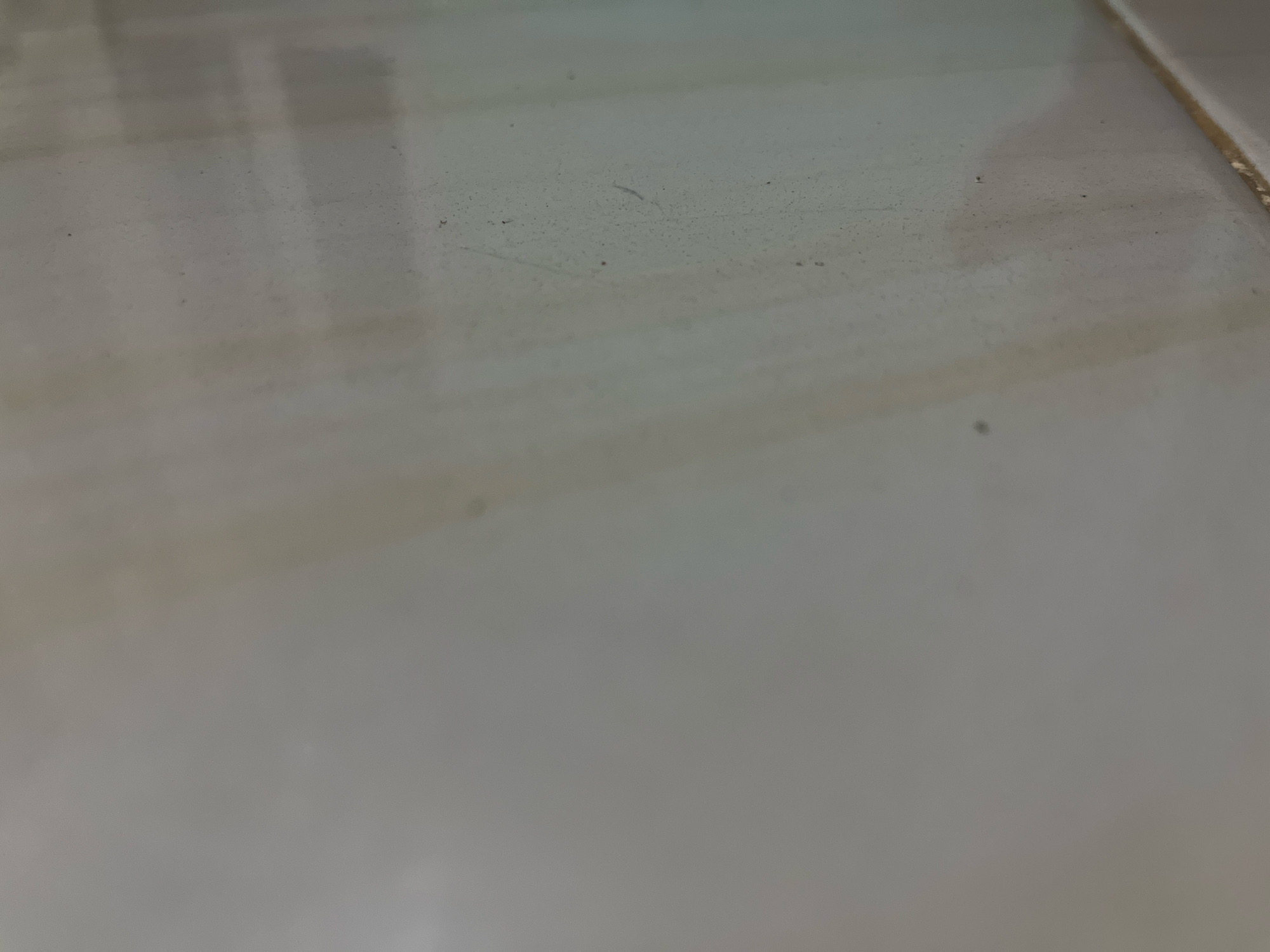 The image shows a close-up view of a tiled floor. The tiles are a light color, possibly white or cream, and appear to be slightly worn or dusty. There are some faint smudges and small dark specks on the surface of the tiles. The lighting in the image is dim, with some areas appearing brighter due to reflections. The perspective is from a low angle, looking across the floor. The overall impression is of an indoor space with a somewhat utilitarian or aged aesthetic. It is not possible to determine the exact location within the city of Kebumen, Indonesia, from this image alone.