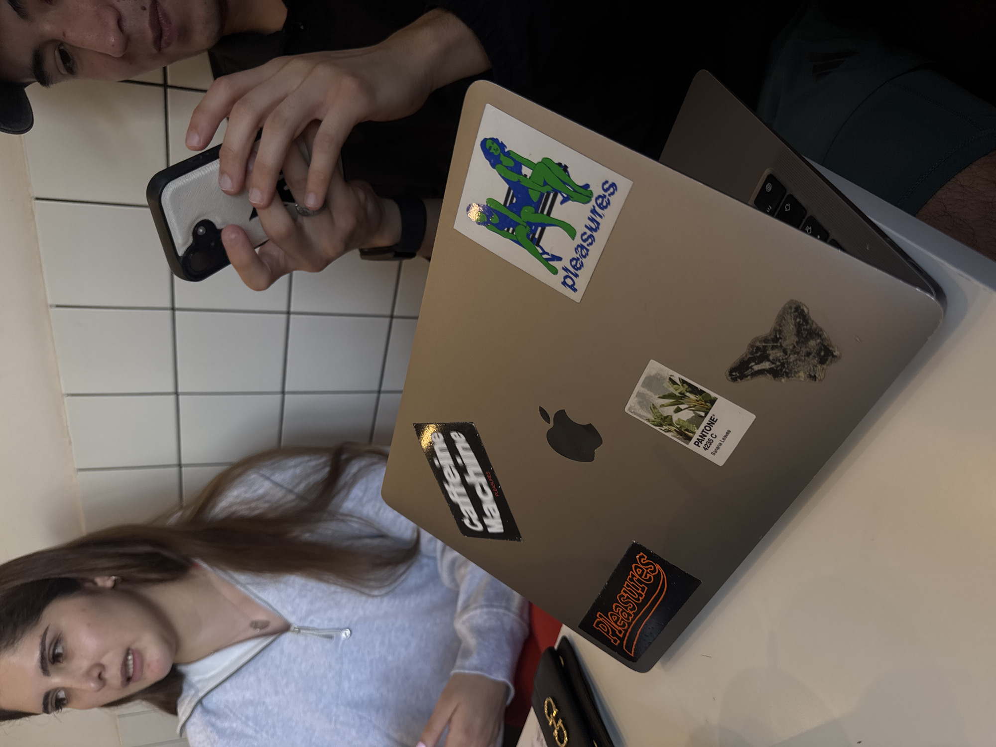 This image depicts an indoor scene, likely a casual setting such as a cafe, in Puebla, Mexico. The perspective is a tilted, downward shot, capturing two individuals and a laptop on a white tabletop.
In the upper portion, a man with short dark hair, wearing a dark top and possibly a cap, is visible from the chest up. He holds a smartphone with a distinctive black and white case in both hands, appearing to interact with it. Behind him is a white tiled wall.
In the foreground, a woman with long brown hair is visible, also from the chest up, wearing a light grey zip-up top. She is looking down and to her left, with a neutral expression.
Between the two individuals on the table is an open, silver MacBook laptop. The laptop lid is decorated with several stickers. Visible text on these stickers includes a blue sticker featuring two green, reclining figures and the word "Pleasures," a black rectangular sticker stating "Caffeine Machine" with smaller "pleasures" text, another black sticker with "Pleasures" in orange text, and a white sticker displaying foliage with the words "PANTONE 4255 C Banana Leaves." A black case or wallet with a gold emblem is partially visible on the table near the woman. The overall atmosphere suggests a casual gathering or work session.