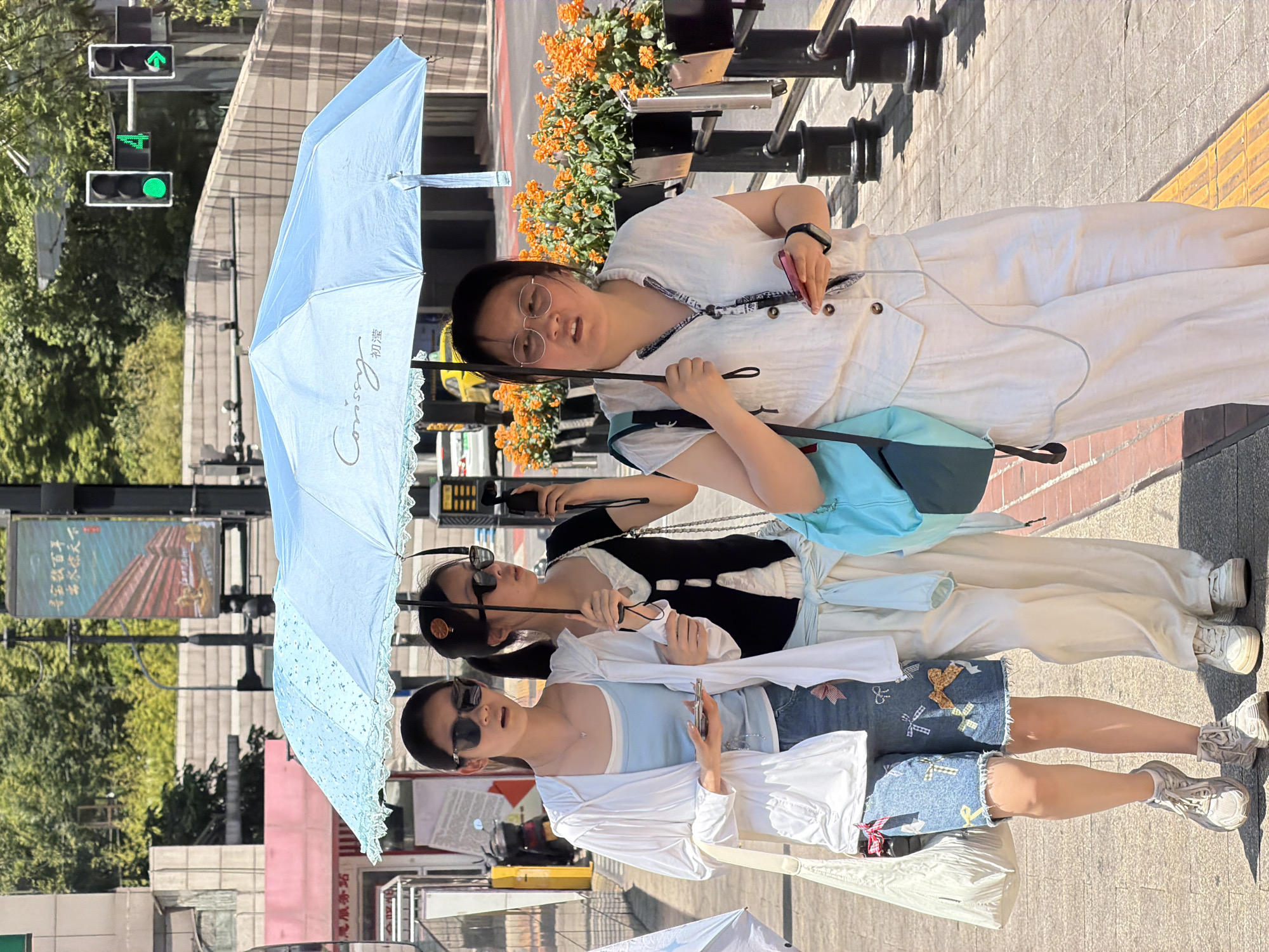 Three young women are walking on a sidewalk under a light blue umbrella, in Yuzhong District, China. The woman on the left wears a white hooded jacket over a light blue tank top and denim shorts adorned with colorful bows. She has sunglasses on and holds a smartphone. The woman in the middle wears a black vest over a white top and cream-colored pants. She has sunglasses on and a hair clip shaped like an orange slice. The woman on the right wears a white button-down shirt and matching cream-colored pants. She wears glasses and holds a pink smartphone and the umbrella's handle. She is looking forward with a neutral expression. The sidewalk is paved with gray and yellow bricks, and there are planters with orange flowers along the right side of the frame. In the background, there are traffic lights, a street pole with an advertisement, buildings, and trees. The scene appears to be a sunny day in an urban environment.