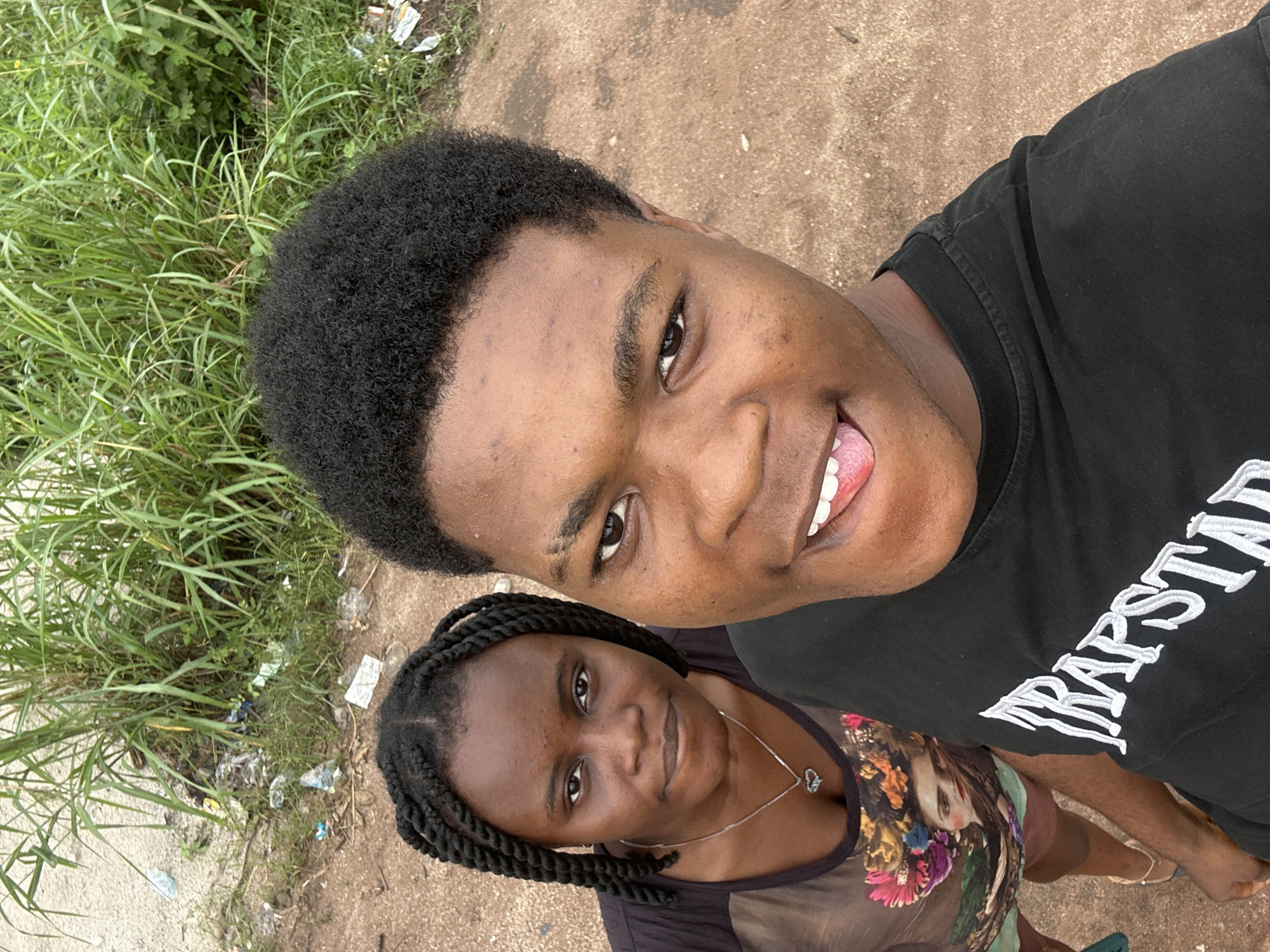 Two individuals, a male and a female, are captured in a close-up, high-angle selfie. The male, positioned closer to the camera and slightly to the right, has dark skin, short, dark, curly hair, and is wearing a black t-shirt with white lettering that appears to read "TRAPSTAR". He is looking directly at the camera with his tongue playfully extended. The female, to the left and slightly behind the male, also has dark skin and her hair is styled in two thick braids. She is wearing a dark, sheer t-shirt adorned with a colorful floral print and a portrait of a woman. A silver necklace with a heart-shaped pendant is visible around her neck. They are standing outdoors on a dirt path. In the background, tall green grass and some scattered litter are visible, suggesting an unkempt environment. The ground is a mix of sand and dirt. The presence of litter and the natural vegetation point towards a location that might be on the outskirts of a more developed area, possibly in a less maintained part of Ihiagwa, Nigeria.