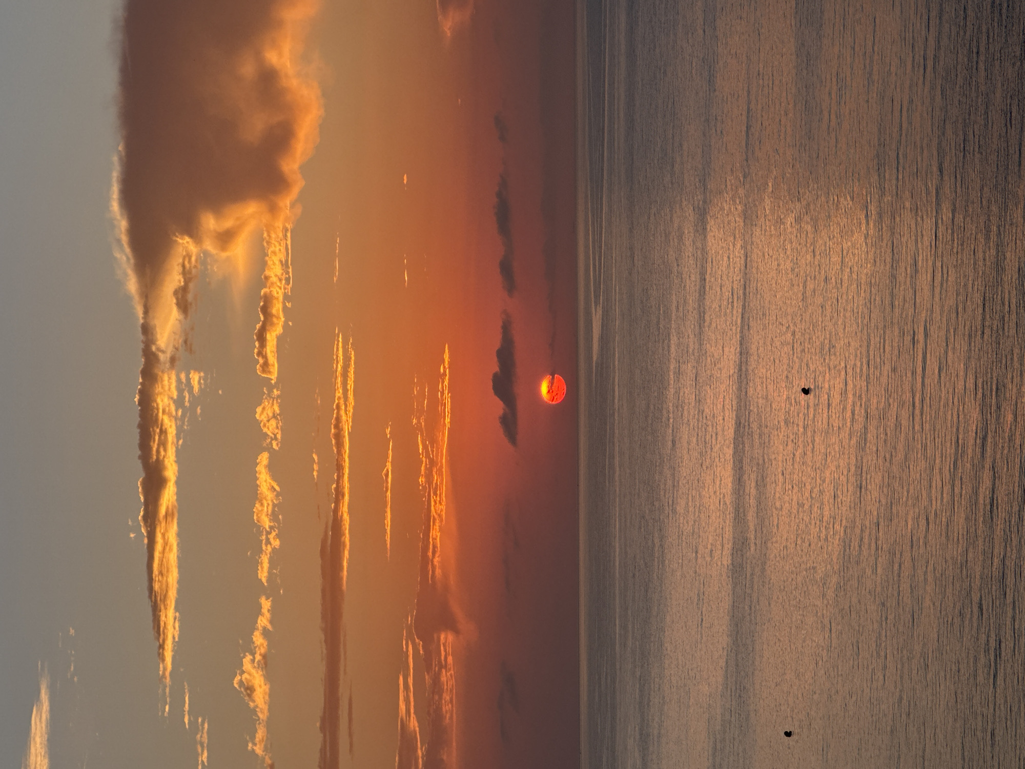 The image depicts a sunset over the Mediterranean Sea, viewed from a high vantage point in Calp, Spain. The sky is ablaze with vibrant hues of orange and red, with scattered clouds catching the last rays of the sun. The sun itself is a fiery red orb, partially obscured by a thin layer of clouds near the horizon. The calm sea reflects the warm colors of the sky, creating a shimmering, golden surface. Two small, dark objects, possibly buoys or small boats, are visible on the water. The overall scene is one of serene beauty, capturing the tranquil end of a day by the coast.
