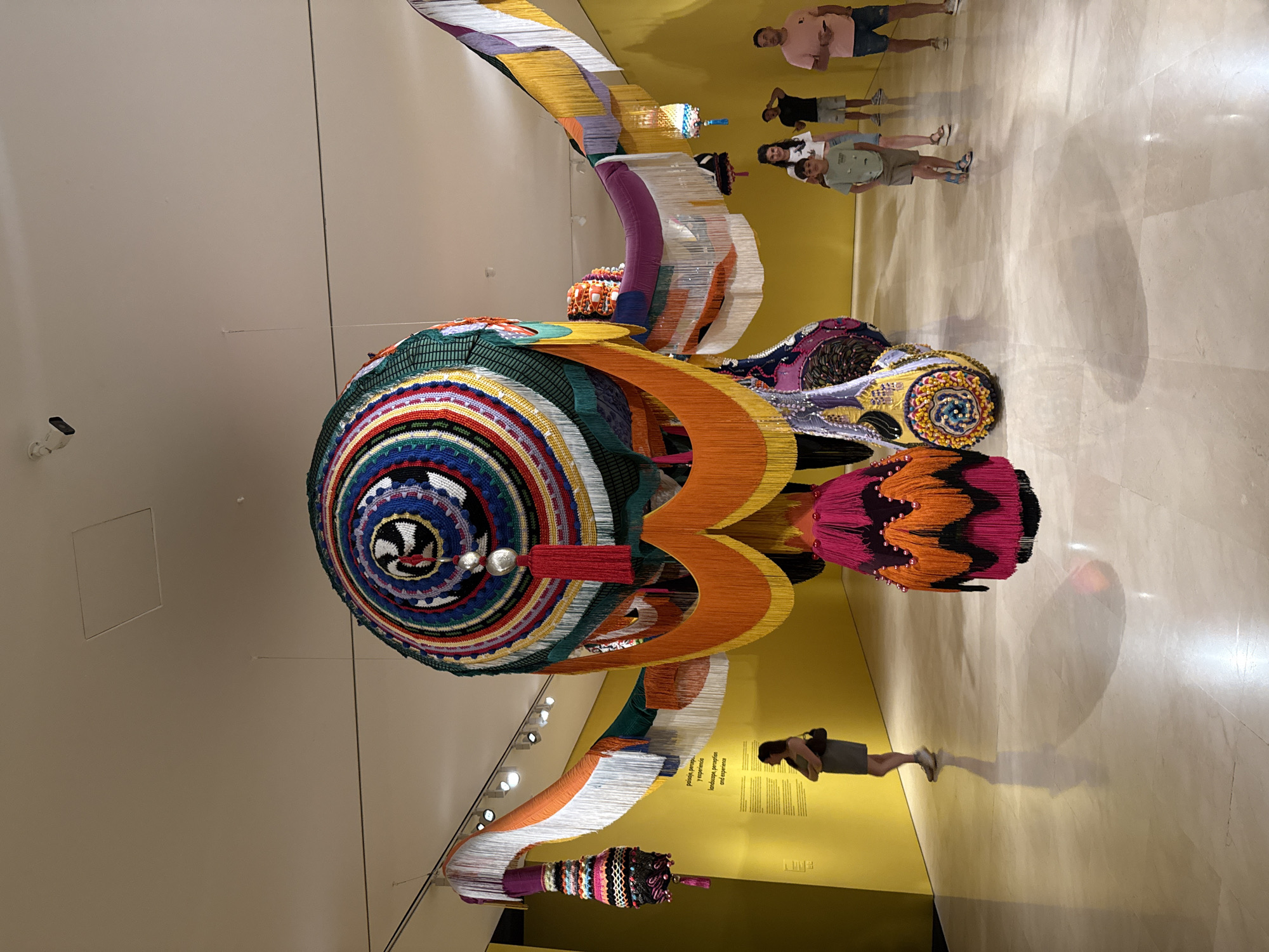 The image depicts a large, colorful, crocheted textile sculpture hanging from the ceiling in a museum gallery. The sculpture is predominantly made of concentric circles of various colors, with intricate patterns and many hanging fringes and tassels. It appears to be suspended by thin wires. In the background, a bright yellow wall features text in both Spanish and English, and a woman in a gray dress stands looking at the artwork, with her arms crossed. Several other people, including children, are visible in the middle ground, observing the sculpture. The floor is polished marble, reflecting the artwork and the people. The lighting in the gallery is primarily from ceiling-mounted spotlights. The scene is consistent with an exhibition at the Centro Pompidou Málaga, given the distinctive yellow exterior visible in other views of this location and the modern art installations often found there.
