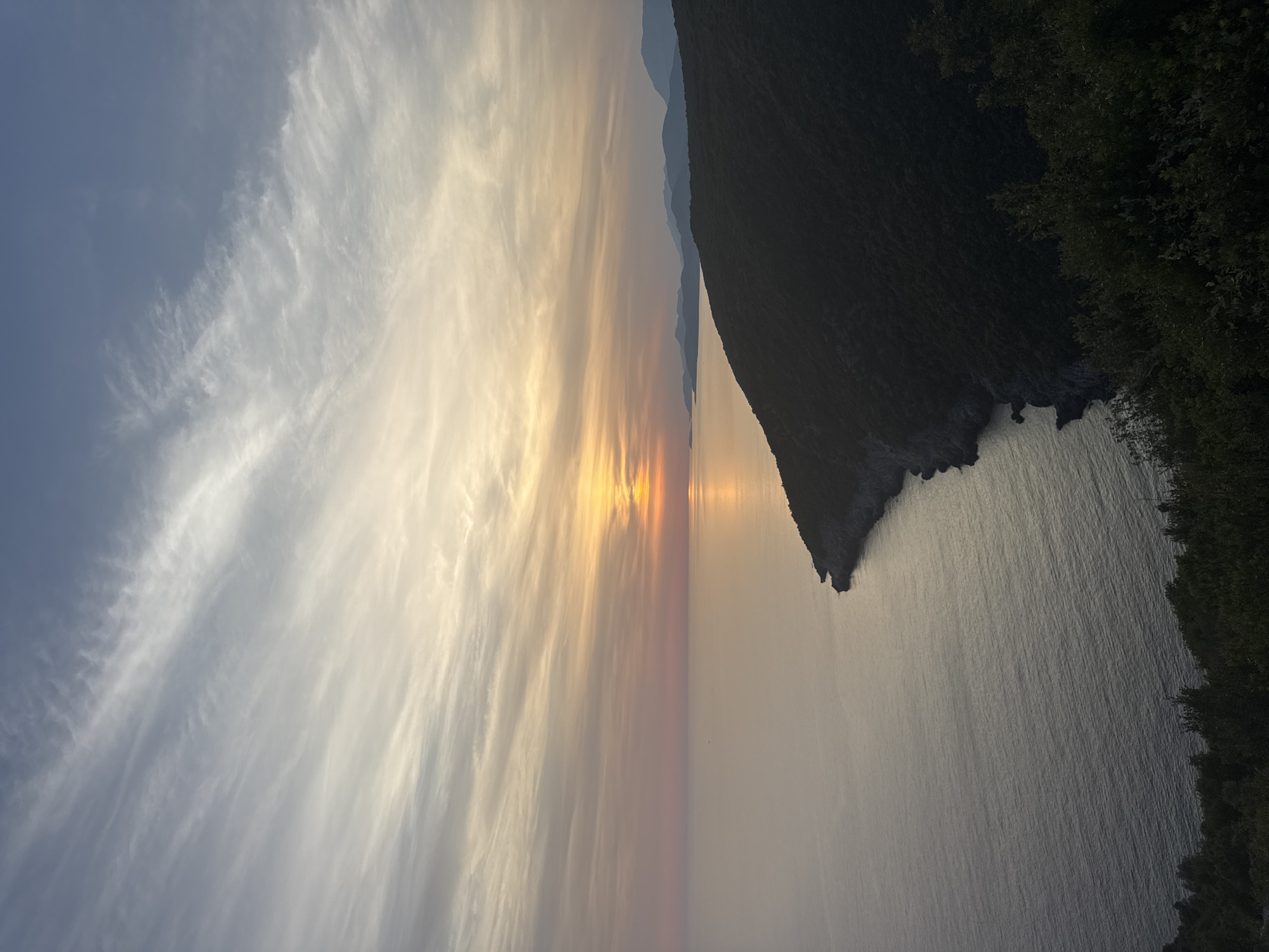 The image depicts a serene coastal landscape at sunset, with the sun casting a warm glow over the Adriatic Sea. The viewpoint is elevated, looking down upon a dark, forested promontory that juts into the water. The sky is a canvas of soft colors, transitioning from pale blue at the top to hues of pink, orange, and yellow near the horizon where the sun is setting. Wispy clouds stretch across the sky, catching the fading light. The sea is calm, reflecting the sky's colors in a muted way, with gentle ripples visible. In the distance, a chain of hazy, silhouetted mountains lines the horizon. The foreground shows the dense green foliage of trees and shrubs, indicating the high vantage point from which the photograph was taken. The overall atmosphere is tranquil and picturesque, characteristic of a coastal sunset. The location is identified as being in the vicinity of Zagora, Montenegro. There are no people or specific structures visible in the image, focusing solely on the natural scenery.