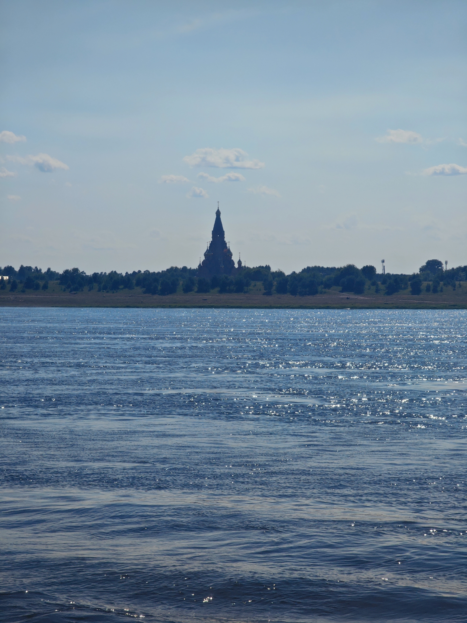 A wide, dark blue river occupies the foreground of the image, its surface rippling with sunlight reflecting off the water. In the midground, a distant treeline of lush green foliage stretches horizontally across the horizon. Silhouetted against a pale blue sky with scattered white clouds, a cluster of buildings with a prominent, spire-like structure stands out on the distant shore. The spire is dark, topped with a cross, suggesting a church or cathedral. The overall impression is one of a serene, expansive landscape on a clear day. The scene depicts the Yenisei River in Lesosibirsk, Russia, with the Transfiguration Cathedral visible in the distance.