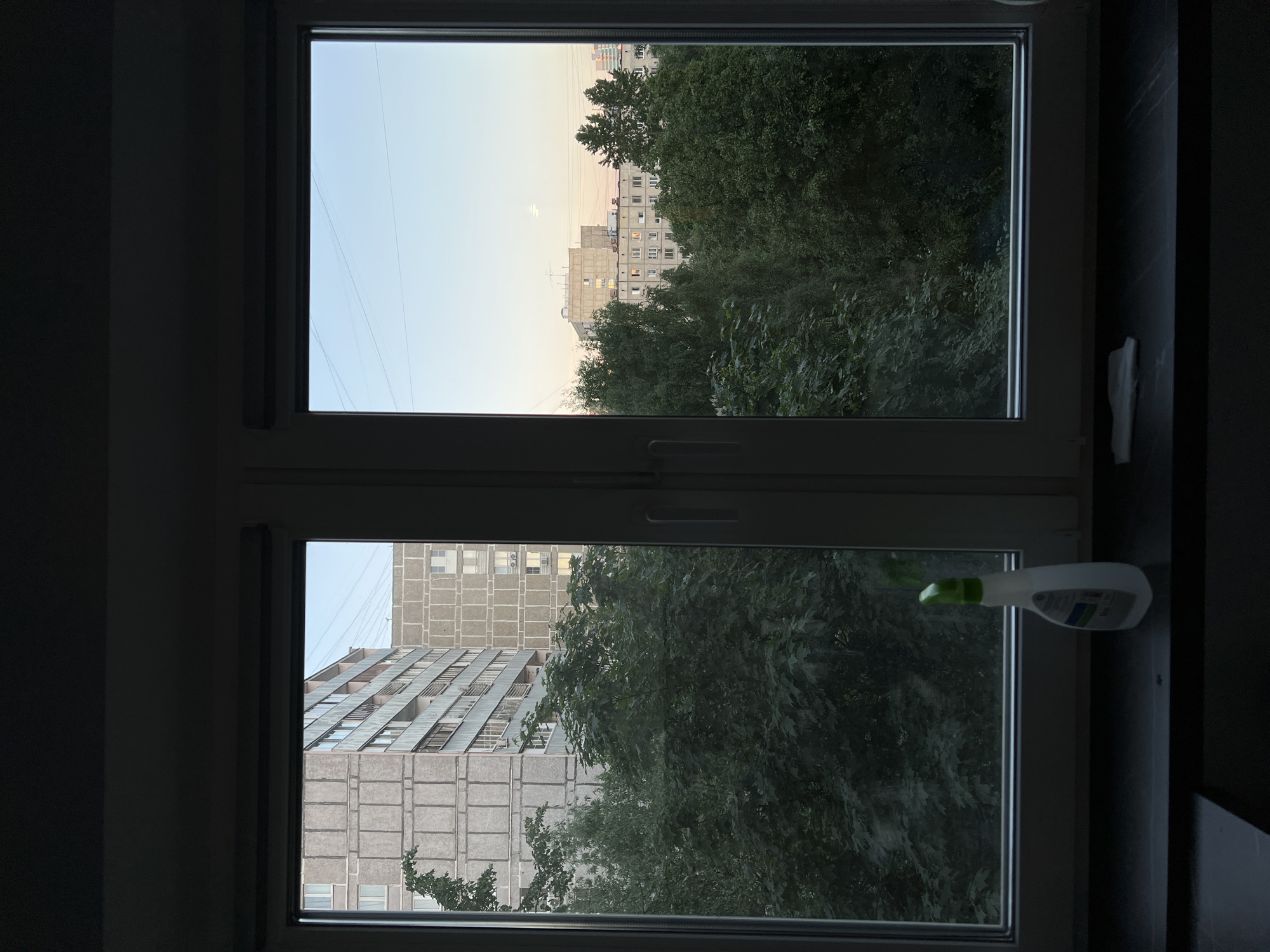 A window in a dim room frames a view of the city of Khimki, Russia. The upper pane shows a pale sky and the tops of trees obscuring a building in the distance, illuminated by the last light of the day. The lower pane gives a closer view of a large, concrete building and foliage. A cleaning bottle is visible on the right window sill. The room itself is dark.