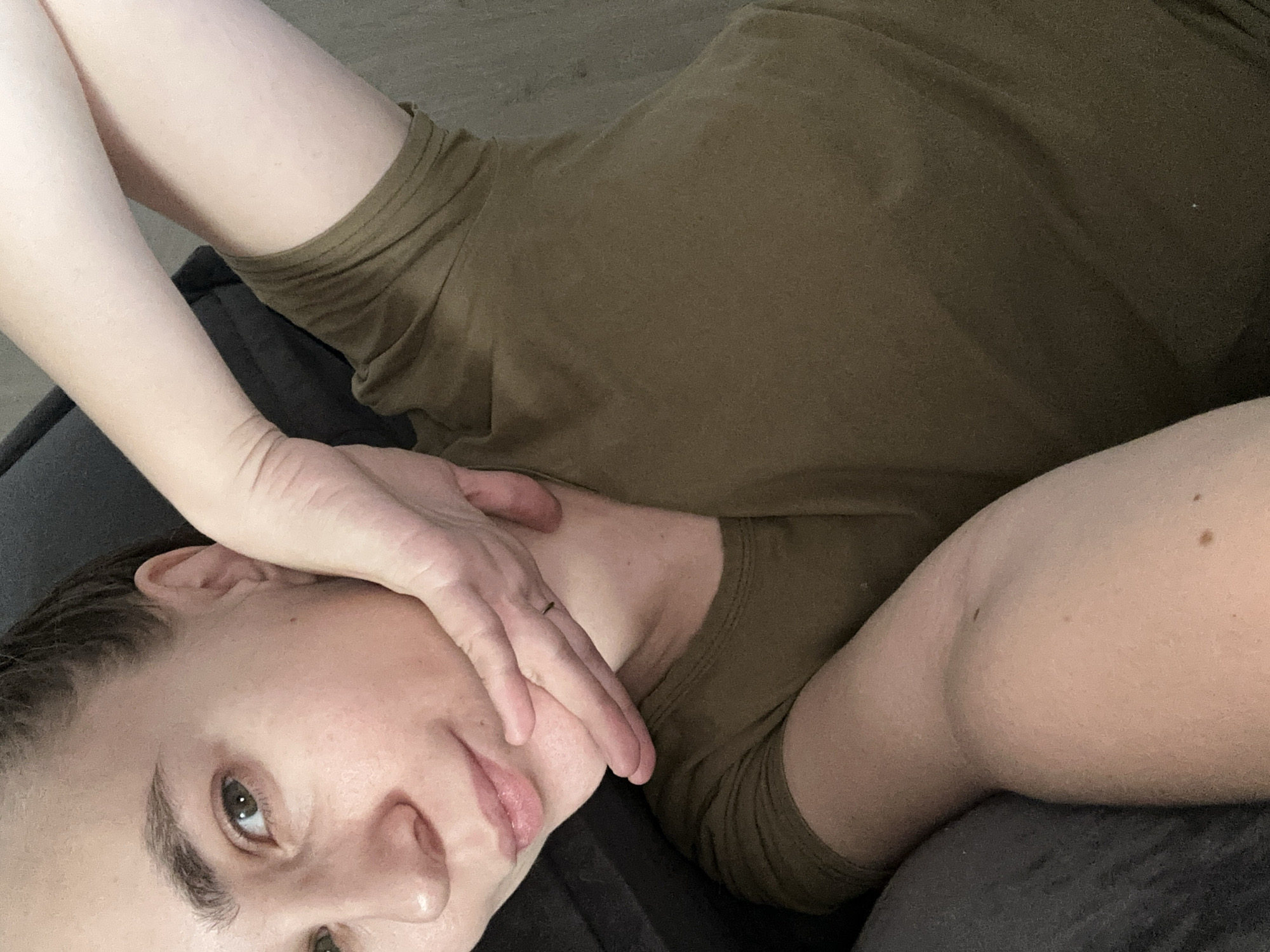 A close-up, slightly overhead shot captures an individual lying down, angled diagonally across the frame. The subject appears to be an adult female, with her head resting on a dark gray or black surface, likely a pillow or cushion. She is wearing a dark olive green, possibly brown, short-sleeved t-shirt. Her right arm is raised, with her hand gently resting against her face, partially obscuring her chin and mouth. A subtle ring is visible on one finger of this hand.
Her eyes, which appear to be a shade of green or hazel, are open and directed towards the viewer, conveying a neutral to slightly pensive expression. Her hair, dark brown, is pulled back, revealing her ear. The lighting is soft and indirect, suggesting an indoor setting. The composition emphasizes the person's face and upper body, with portions of her left arm and torso visible. Skin texture, including a few moles, is visible on her arm and shoulder. The overall impression is one of relaxation or quiet contemplation within a private indoor space, located in Kharnavy, Russia. There is no visible text.