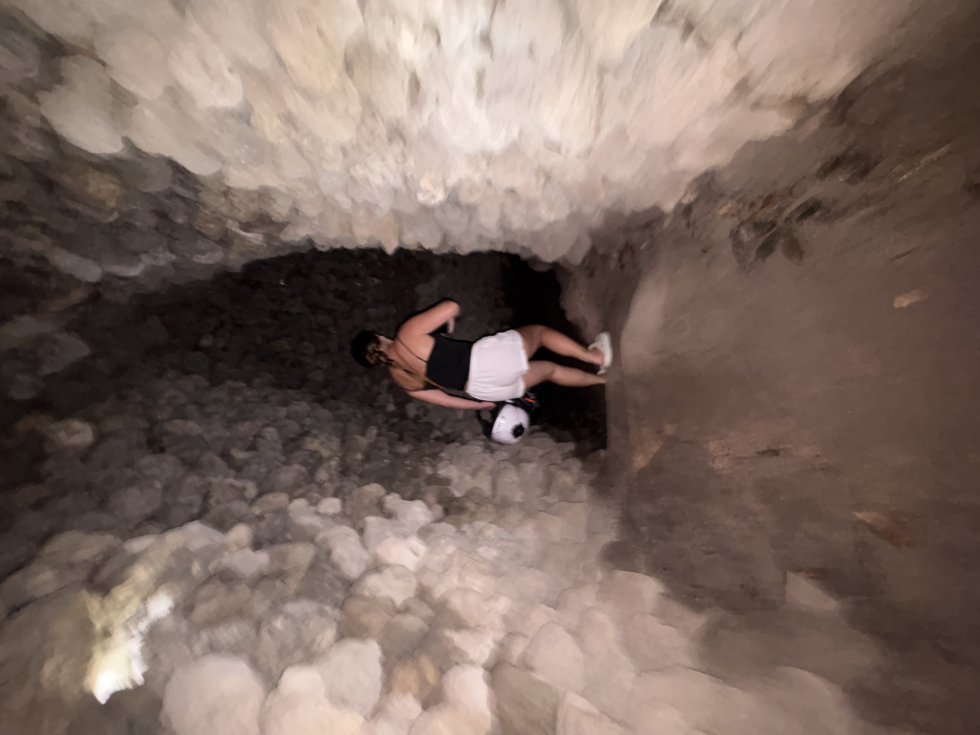 An individual is depicted exploring a narrow, rocky cave passage in Cam Ranh, Vietnam. The scene is dimly lit, typical of a subterranean environment. The person, viewed from behind and slightly to their left, is wearing a black sleeveless top, white shorts, and white sneakers. They are holding a white helmet or a spherical object with an integrated light source, which casts a bright beam onto the uneven, stone-covered cave floor directly in front of them.
The cave walls and ceiling are composed of rough, light-colored rock formations, creating a rugged and enclosed space. The ground is similarly covered with an accumulation of irregular rocks and debris, suggesting challenging terrain for traversal. The person's posture indicates they are carefully navigating or pausing during their exploration. There are no discernible external light sources, and the time of day is irrelevant within the confined cave. No text is visible within the image.