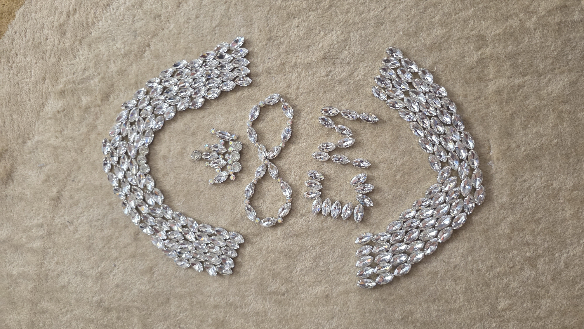 The image displays an overhead view of numerous clear, faceted rhinestones, primarily marquise-shaped, arranged on a textured, light brown or beige soft fabric surface.
Two large, symmetrical formations of rhinestones flank the scene, positioned on the left and right sides. Each formation consists of multiple layers of stones, creating a substantial, curved, wing-like or bracket-like shape that arcs inwards.
In the central area, several smaller arrangements of single-layered rhinestones are visible. A prominent vertical shape clearly resembles a figure-eight or an infinity symbol. To its upper left, a small cluster of rhinestones, some exhibiting an iridescent sheen, forms a star-like or floral pattern. To the upper right of the central figure-eight, a chain of three interconnected pairs of rhinestones creates a shape resembling a stylized '3' or 'S'. Below this, another curved arrangement of rhinestones forms an open circle, similar to a 'C' or '6'.
The deliberate arrangement of the rhinestones suggests a creative activity, possibly related to crafting, costume design, or artistic embellishment. The sparkling quality of the stones against the neutral background evokes a sense of glamour and intricate detail.
Location context: Blagoevgrad, Bulgaria.