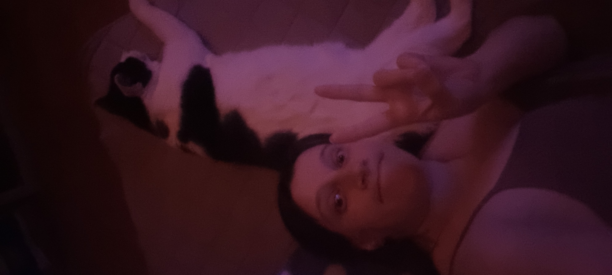 A person is lying down next to a black and white cat. The person is holding up two fingers, making a peace sign. The location is not provided in the image.