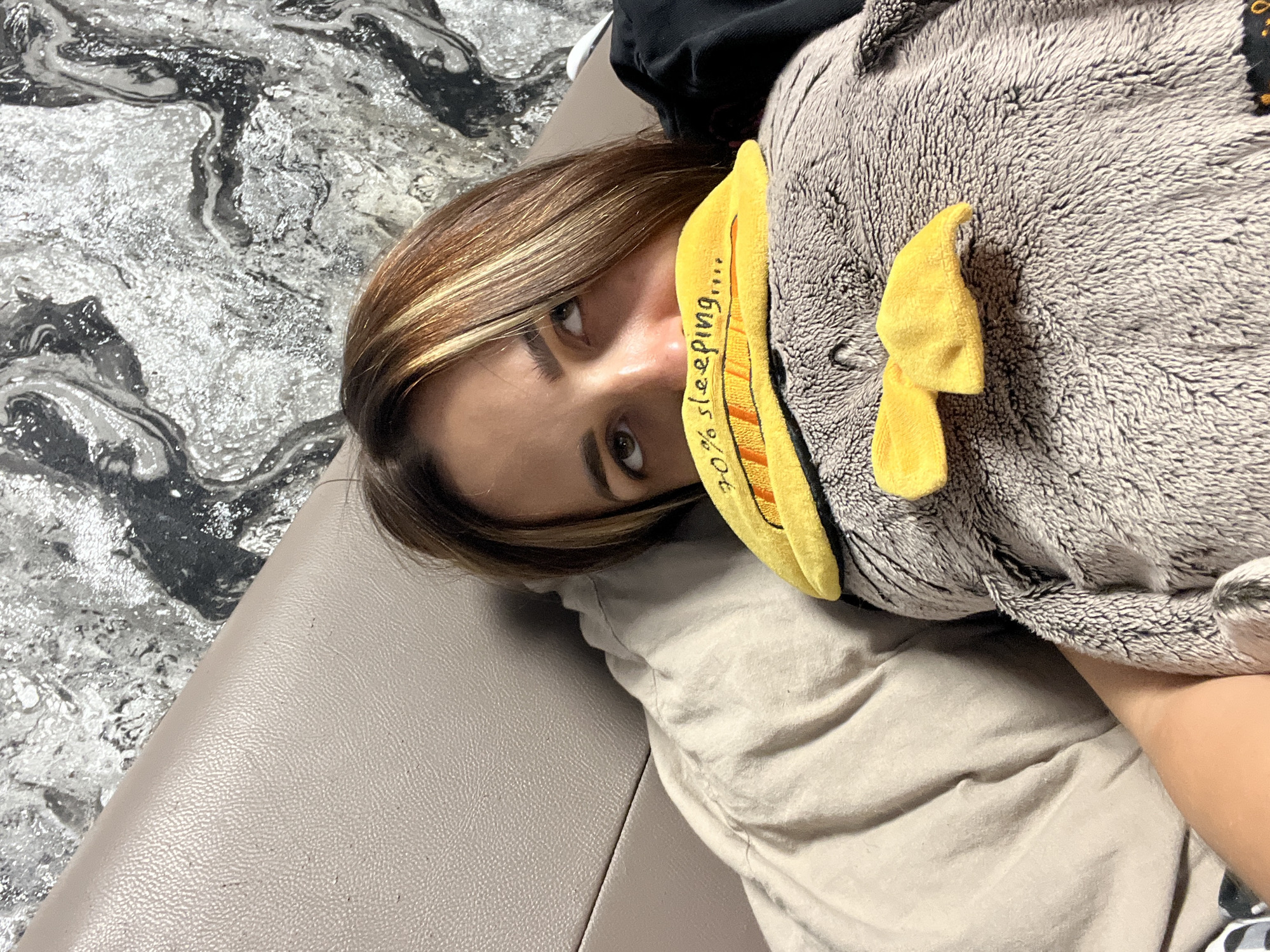 The media file is a high-angle, close-up, portrait-oriented photograph of a person lying down, partially obscured by a stuffed animal. The person has light brown hair with blonde highlights and is looking towards the camera with a neutral expression. Their right eye is clearly visible, with a brown iris. A yellow fabric piece, resembling a beak or a bib, is positioned over the person's mouth and nose. This yellow fabric has black lettering that reads "90% SLEEPING....".
The person is lying on a surface that appears to be a couch or bed, with a textured, light-colored fabric underneath. To the left of the person and the stuffed animal, there is a surface with a marbled pattern of black, white, and silver, possibly a countertop or a decorative surface. The lighting suggests it is indoors, and the time of day is indeterminate. The overall mood of the image is casual and possibly humorous, given the text on the fabric.