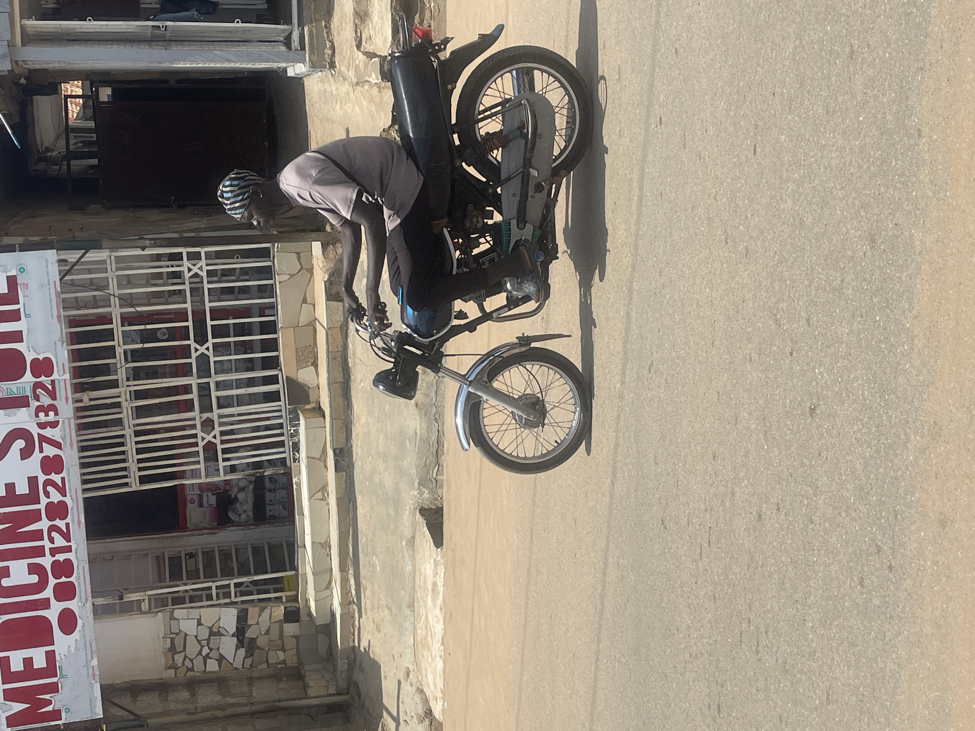 In Abuja, Nigeria, a person is riding a motorcycle down a paved road. The person is wearing a grey t-shirt, black pants, and a headscarf with a blue and white striped pattern. They are operating a black motorcycle with silver accents. Behind the motorcycle rider, a building with a sign that reads "MEDICINE STORE" is visible. The building appears to be a commercial establishment with a metal gate and a stone facade. The scene suggests a typical day on a city street in Abuja.