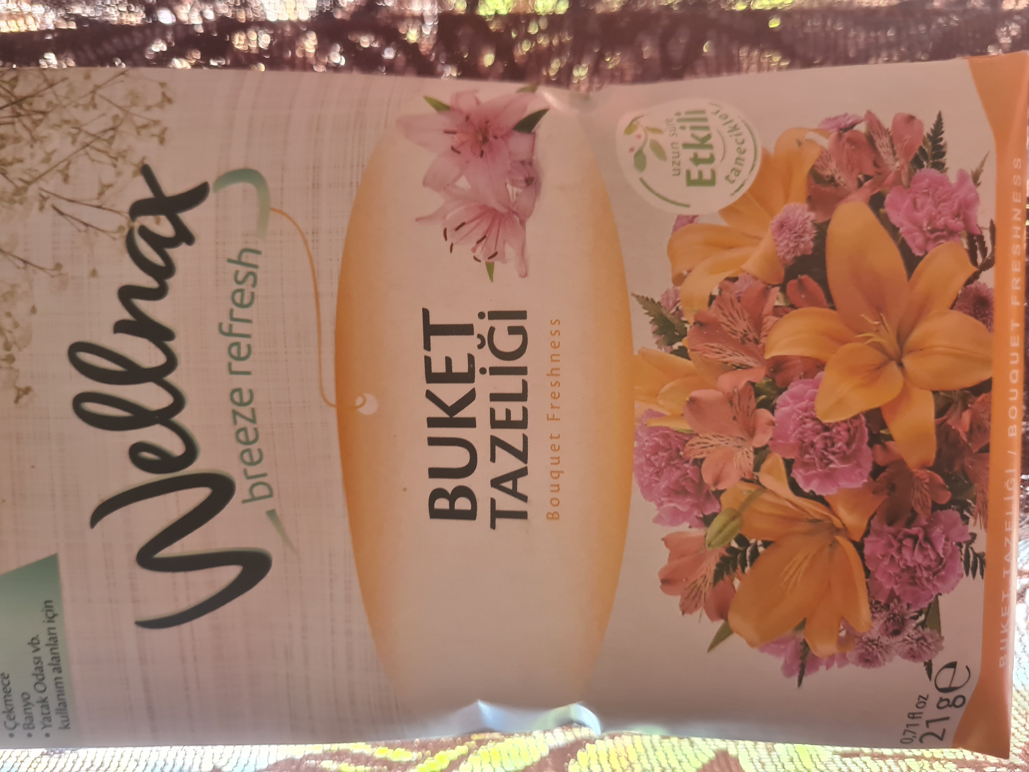 This media file is a close-up of a product packaging. The packaging features a floral design with large orange lilies and smaller pink flowers. Prominently displayed is the brand name "Wellmax" in large, cursive black lettering, accompanied by the text "breeze refresh". Below this, in vertical orientation, is "BUKET TAZELİĞİ" and "Bouquet Freshness". To the left of the floral arrangement, there is a green circle with Turkish text which translates to "Long-lasting Effective Scents". On the bottom left, there is a list of suggested uses in Turkish: "Çekmece" (Drawer), "Banyo" (Bathroom), and "Yatak Odası vb. kullanım alanları için" (for areas like bedrooms, etc.). At the bottom right, there are product specifications: "0.71 fl oz" and "21 ge". The packaging appears to be for an air freshener or a similar home fragrance product. The background is a textured, sparkly fabric, possibly a tablecloth or a decorative surface, suggesting the product is displayed for promotional purposes. There is no indication of people or a specific time of day or weather. The text is in both Turkish and English.