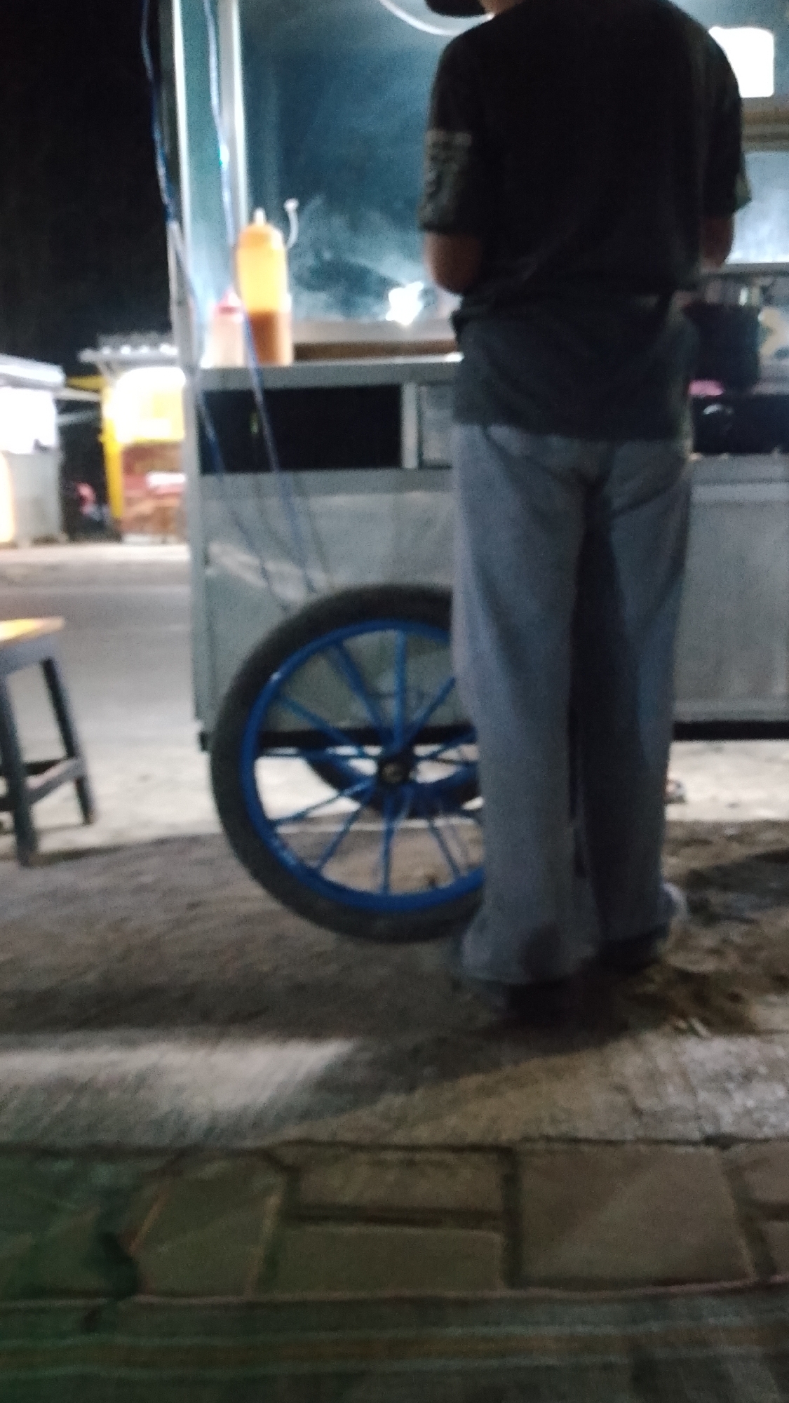 In Karanglangu, Indonesia, a person is standing near a food cart. The individual, viewed from behind, is wearing a dark t-shirt and light-colored pants. They are positioned to the right of the food cart, which has a prominent blue-rimmed wheel. A yellow squeeze bottle is visible on the cart's surface, along with other indistinct items. The ground beneath the cart and person appears to be dirt and paving stones. The background is dark, suggesting it is nighttime or a poorly lit area.