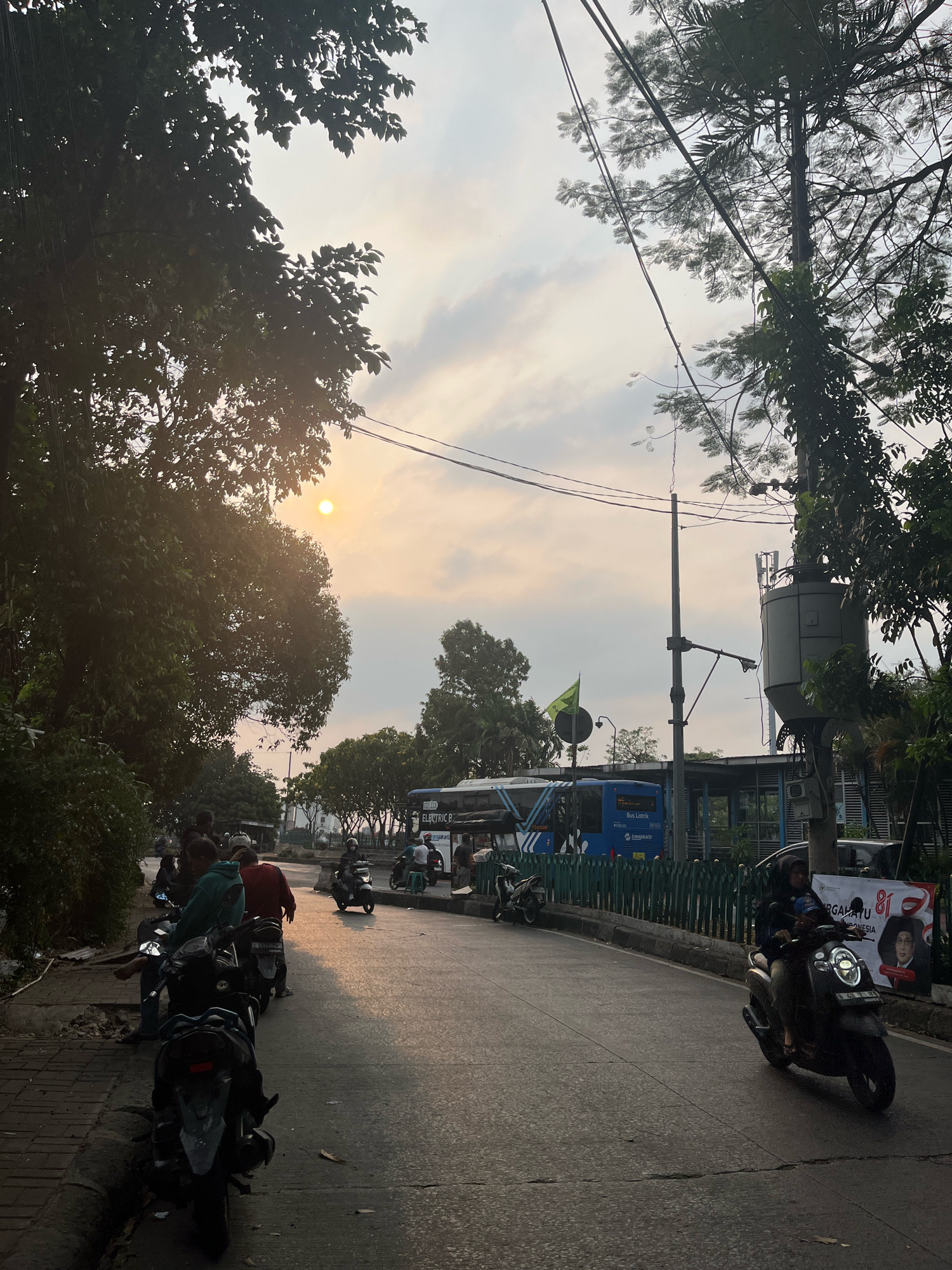 The image captures a street scene in East Jakarta, Indonesia, during what appears to be late afternoon or early evening. The setting sun, a bright orb in a hazy sky, casts a warm glow and creates long shadows across the asphalt road. The left side of the road is lined with trees, their leaves silhouetted against the sky, and a sidewalk with several parked motorcycles. A group of people are gathered near the motorcycles, some standing and others seated.
The road itself is paved and shows signs of wear. A green metal fence separates the road from a raised area where a blue TransJakarta bus is visible, with the words "BUS LISTRIK" and "TransJakarta" printed on its side, indicating it is an electric bus. Further down the road, more motorcycles are seen, some with riders. On the right side of the road, a tall utility pole with cables and a transformer is prominent, along with dense foliage from trees. A banner with Indonesian text and a portrait of a man is also visible on the right. The overall atmosphere is one of urban activity in a tropical environment.