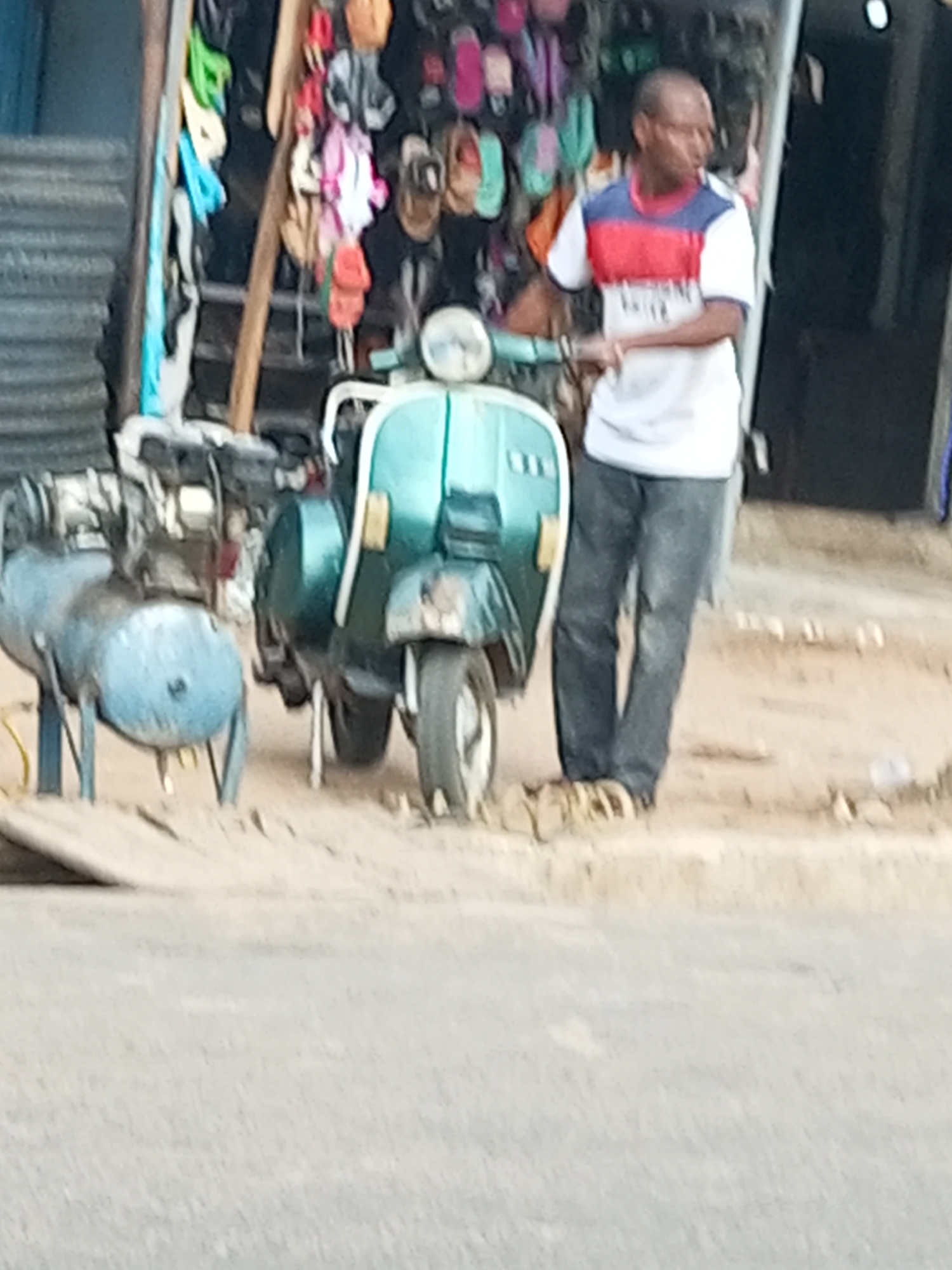 The image depicts a street scene in Arawa, Nigeria. In the foreground, a light blue air compressor is positioned on the ground. To its right, a teal-colored scooter is parked. A man wearing a red, white, and blue striped t-shirt and dark pants stands next to the scooter, facing away from the viewer. Behind the man and scooter, a stall displays various colorful flip-flops and sandals. A corrugated metal structure is visible to the left of the stall. The ground appears to be dirt or sand.