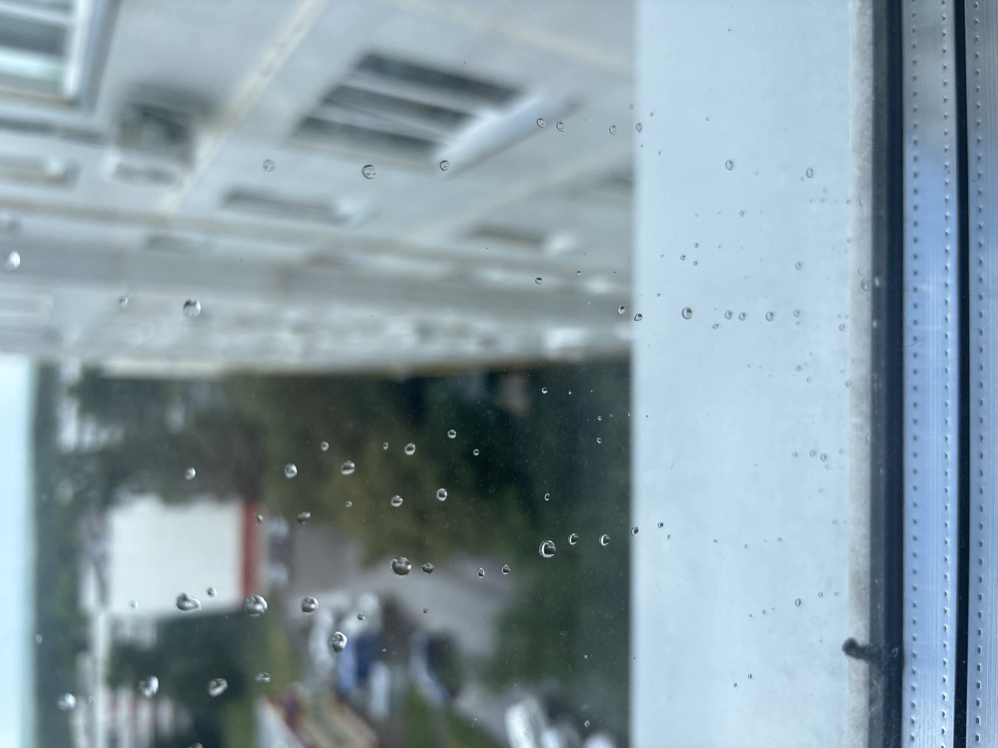 The image depicts a close-up view of a window with raindrops on its surface. Through the raindrops and the slightly blurred glass, a city scene is visible. In the background, there is a multi-story building with visible windows and an air conditioning unit. To the left of the building, there appears to be a street with parked cars and other buildings, suggesting an urban environment. The overall lighting suggests it is daytime, possibly overcast or raining, consistent with the presence of raindrops on the window. The foreground shows the window frame, a metallic structure with a textured surface. The location is identified as Naberezhnye Chelny, Russia.
