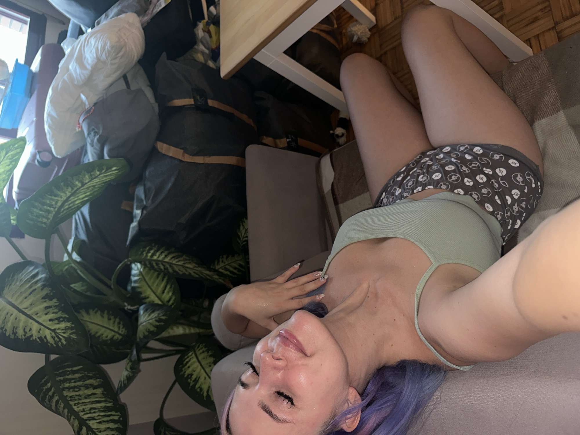 A female-presenting individual with purple hair, closed eyes, and a slight smile is depicted in a close-up, upward-angle selfie. She wears a light green tank top and dark gray shorts patterned with white and light gray circular motifs. Her left hand, featuring manicured nails with embellishments, rests near her neck, displaying subtle glitter on her arm and shoulder. She is lying on a light gray upholstered furniture piece, partially covered by a brown and beige patterned blanket.
The background reveals an indoor setting. To the left and upper-middle, a large plant with variegated green leaves is prominent. Behind it, a stack of items includes multiple large, dark-colored duffel bags with light brown straps, a rolled white item, a reddish-brown container, and a blue plastic container. On the far right, a light-colored wooden table with white legs is visible. The floor beneath the table is wooden with a parquet pattern. Natural light appears to enter from a window in the upper background. The scene is situated in Gijón/xixón, Spain.