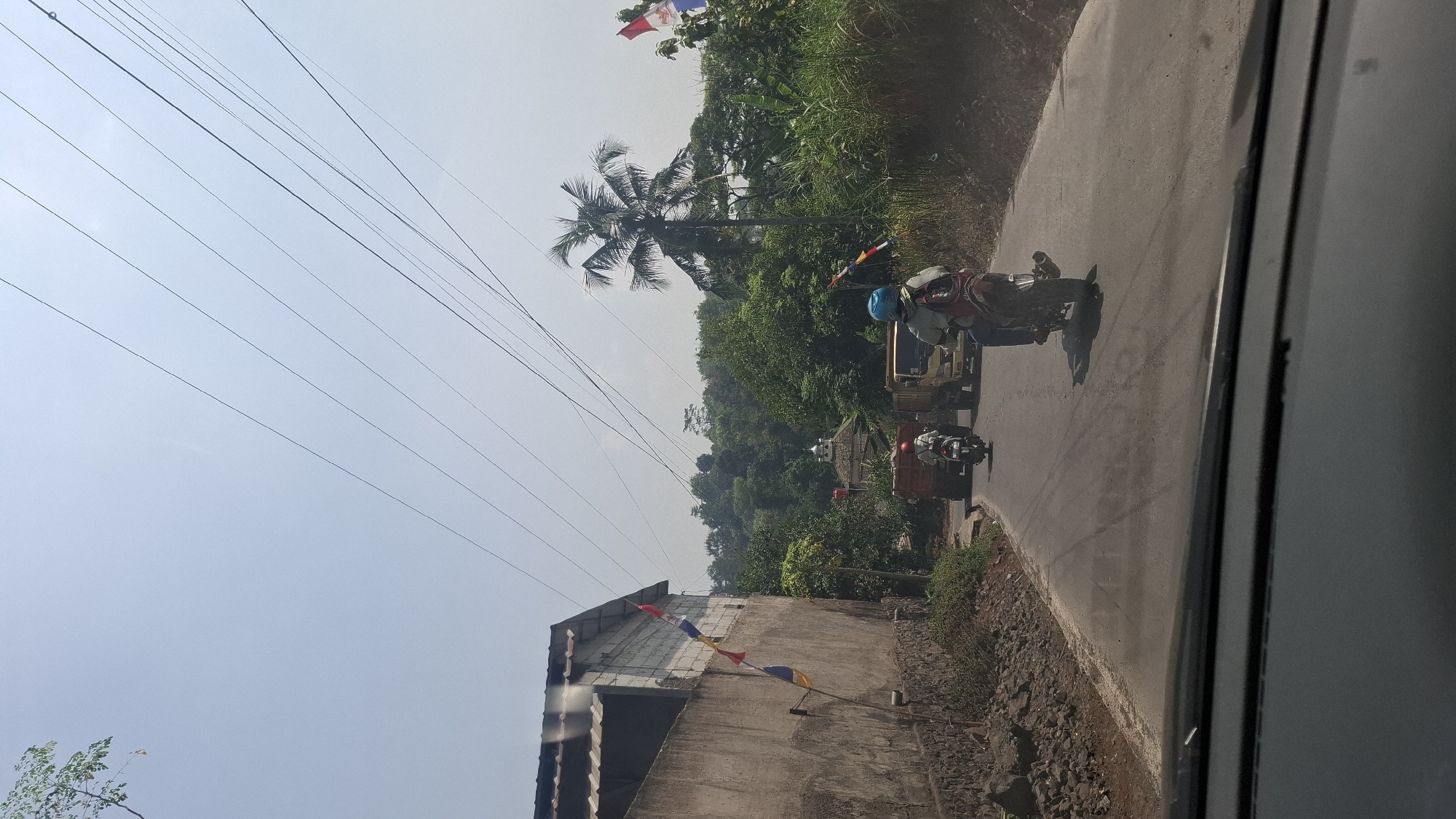 The image depicts a road in Sirnagalih, Indonesia, on a sunny day, with power lines stretching across the sky. On the left, a stone wall with a rough texture runs along the side of the road. A building with a corrugated metal roof and white cinder block walls is situated behind the wall. Several small, colorful flags are attached to the building and the wall.
The road is asphalt and appears to be a two-lane road. Several vehicles are visible on the road. In the foreground, from the perspective of a vehicle's interior, the top edge of a dashboard is visible. Ahead on the road, two motorcycles are traveling in the same direction. The rider of the rear motorcycle is wearing a helmet and a backpack. Further ahead, a small truck and a larger truck are also on the road.
The environment surrounding the road is lush and green, with dense trees and foliage. A tall palm tree stands prominently in the middle ground. In the background, a building with a domed roof, possibly a mosque, is visible amidst the trees. The sky is clear and bright blue. The overall impression is a rural or semi-rural road with moderate traffic.
