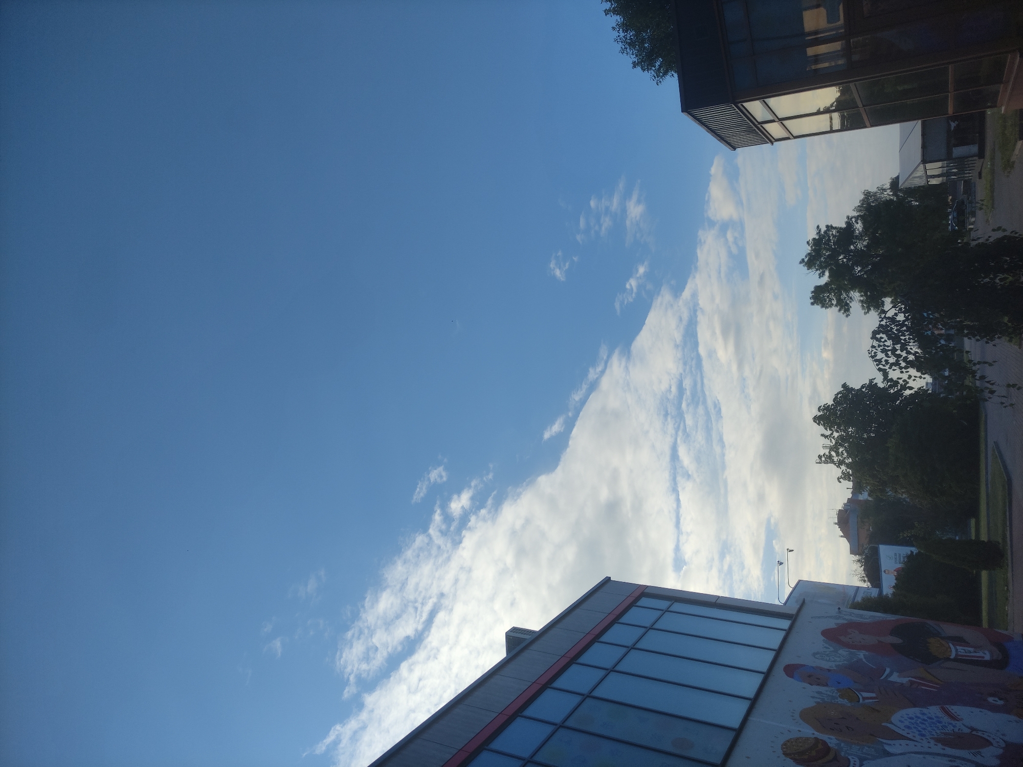 The image depicts an outdoor scene in the city of Mahilyow, Belarus, likely during daylight hours with a clear sky. In the foreground, to the left, is the side of a modern building with large glass windows and a section of a mural featuring stylized figures. To the right, another modern building with reflective glass windows is visible. Trees and other structures, including some buildings in the distance, are present in the middle ground and background. The sky is predominantly blue with scattered white clouds. The perspective is from ground level, looking upwards and across the cityscape.