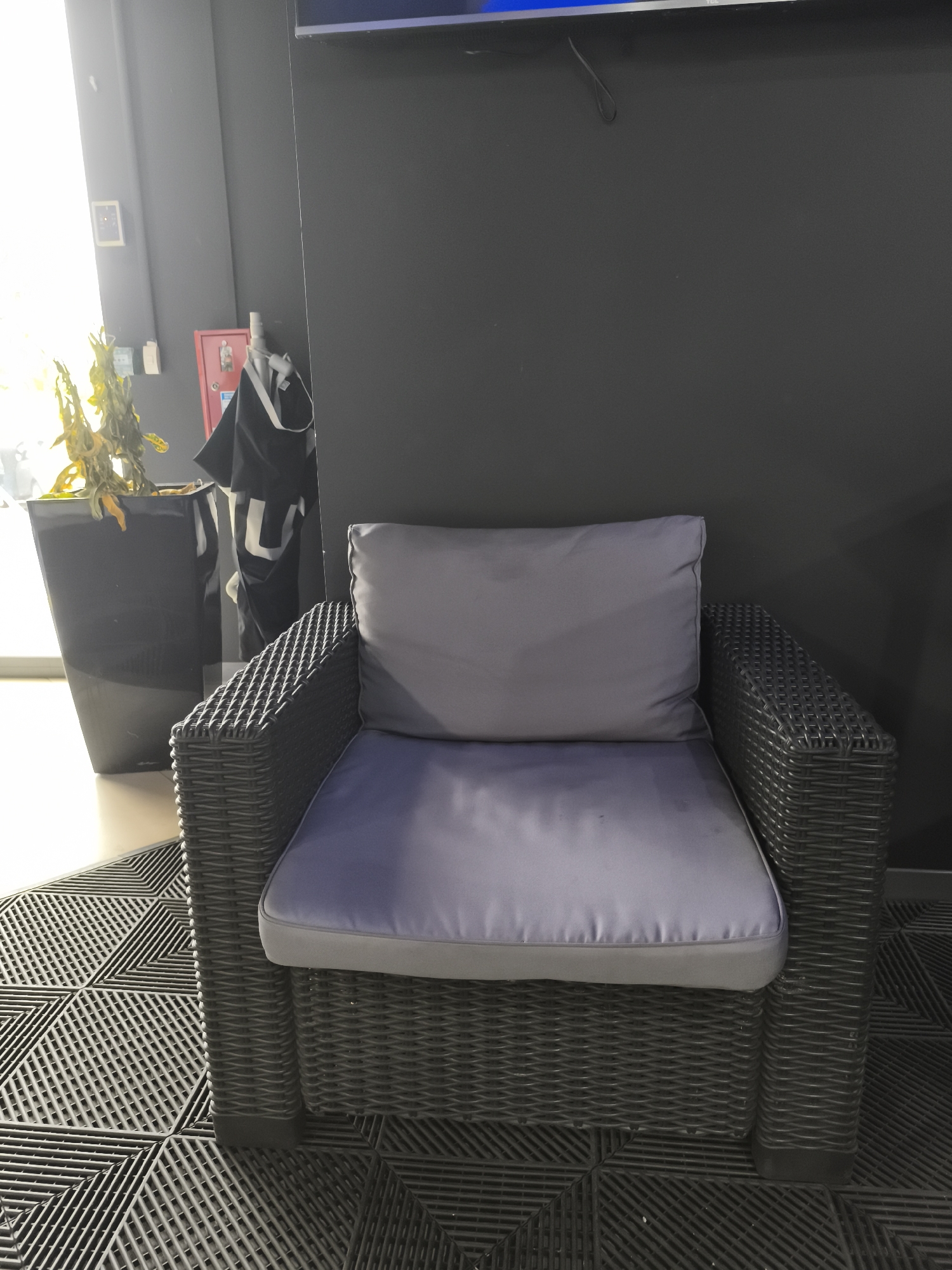 A dark grey, wicker-style armchair with grey cushions is positioned on a black, interlocking tile floor. Behind the chair, a dark grey wall serves as a backdrop. To the left of the armchair, a tall black planter holds a wilting, yellow-leafed plant. Further left, a bright window allows natural light to illuminate the scene. A red fire alarm box is mounted on the wall, with a black garment hanging from it. The environment appears to be indoors, possibly a commercial or communal space in Saint-Denis, France. The image is taken from a slightly low angle, focusing on the armchair and its immediate surroundings.